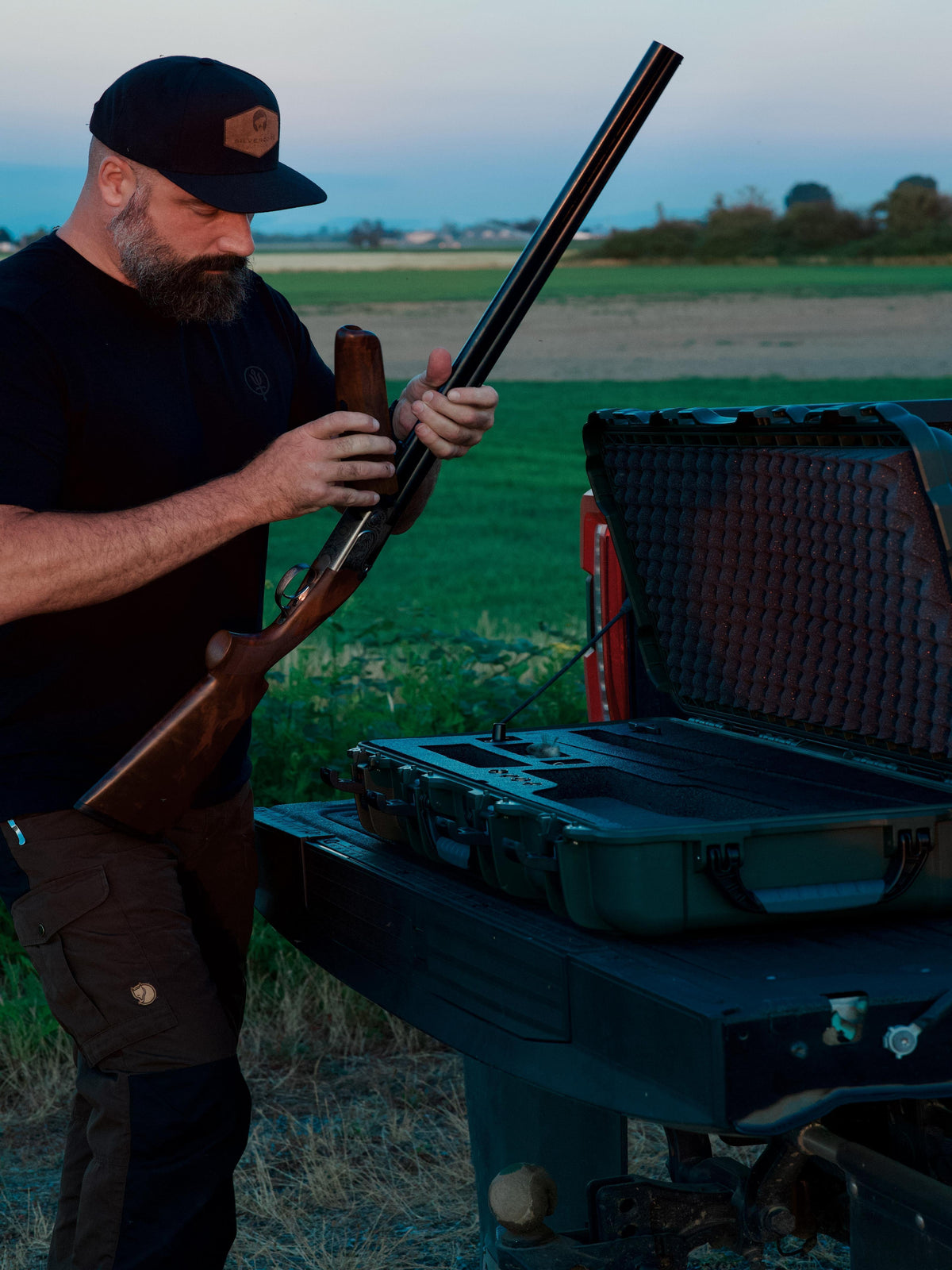 A bearded man in a black cap and black shirt holds a long gun with a wooden stock next to an open black hard case on a table in an outdoor field at dusk.