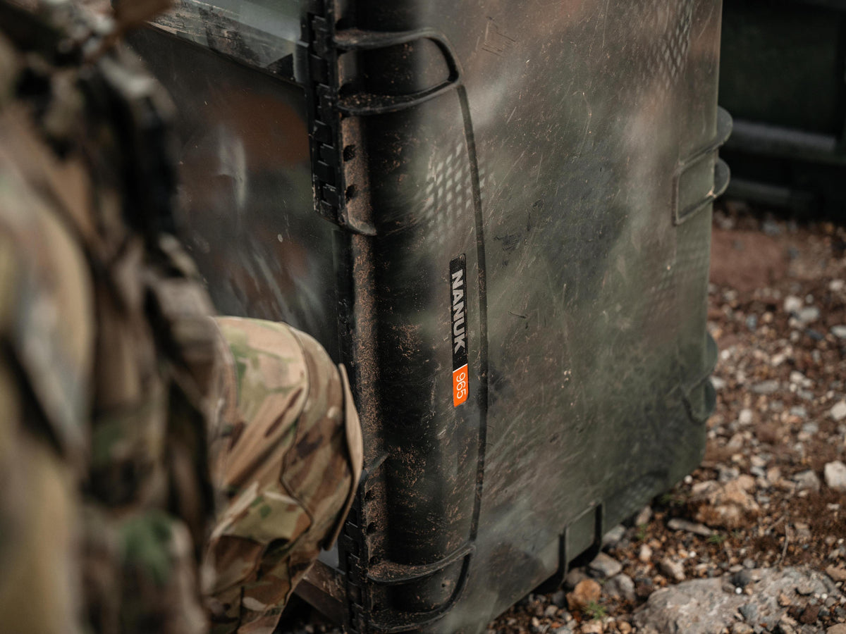 Close-up of a black tactical hard case with a camouflage-patterned strap and a visible NANGUK brand label, shown outdoors near rocky ground.
