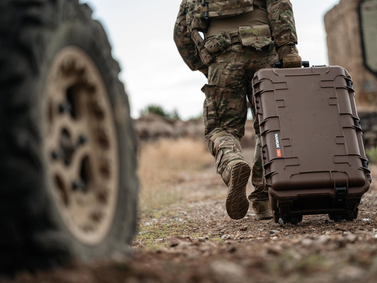 A person in camouflage military uniform walks outdoors while pulling a brown hard-sided rolling case with wheels. The scene is outdoors on a dirt path with a large blurred tire in the foreground and a vehicle or structure in the background.