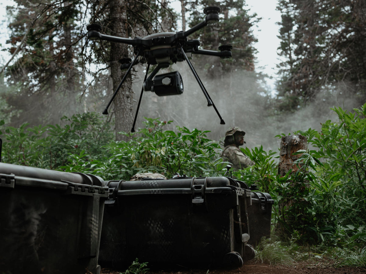 A black quadcopter drone with a mounted camera hovers over a forest clearing above two soldiers standing behind dark hard cases. Dense green foliage and trees fill the background, with mist or dust in the air.