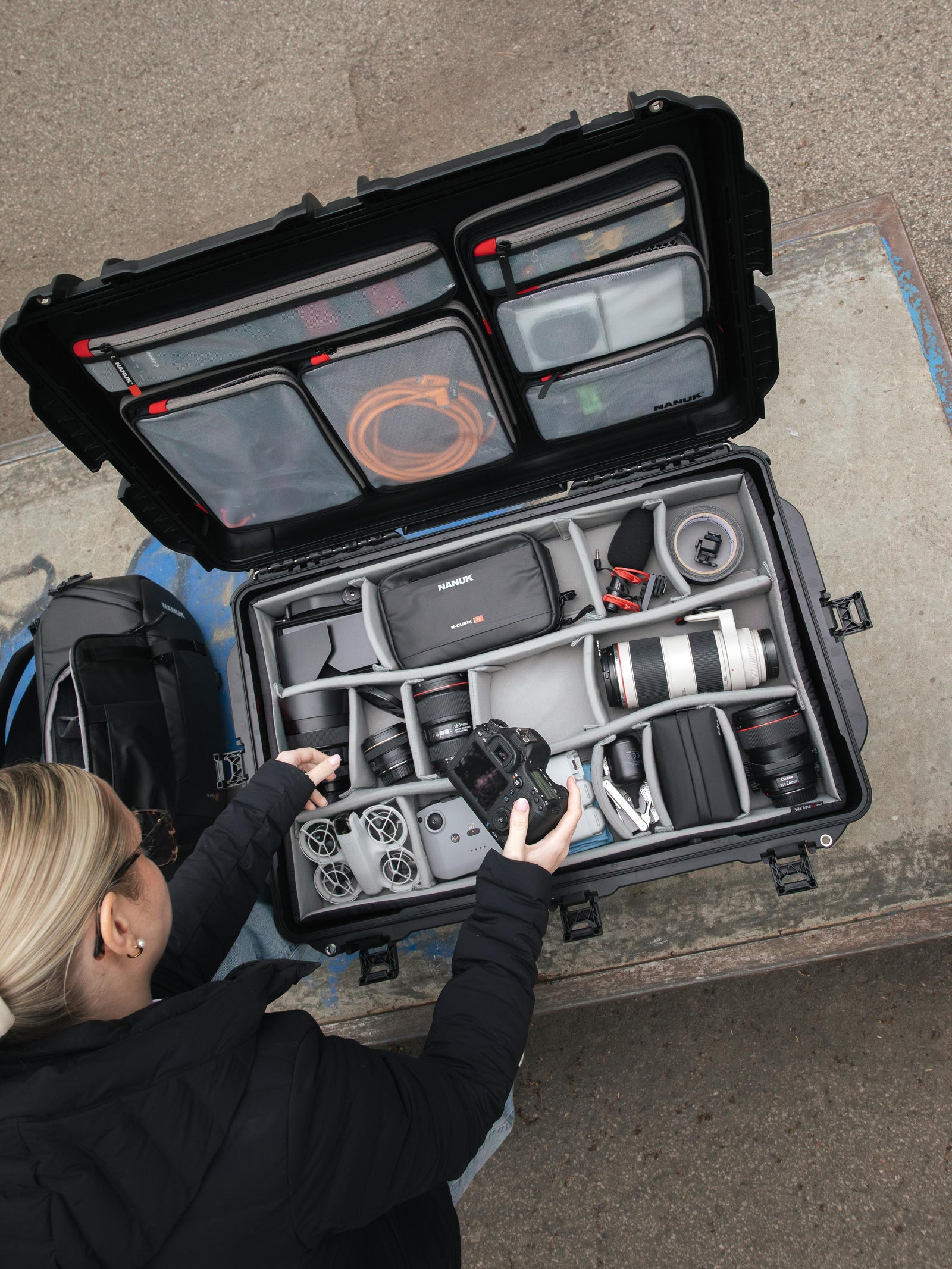 An open black hard-shell camera gear case on a concrete surface, filled with lenses, accessories, and a camera, with a person reaching in from the left.