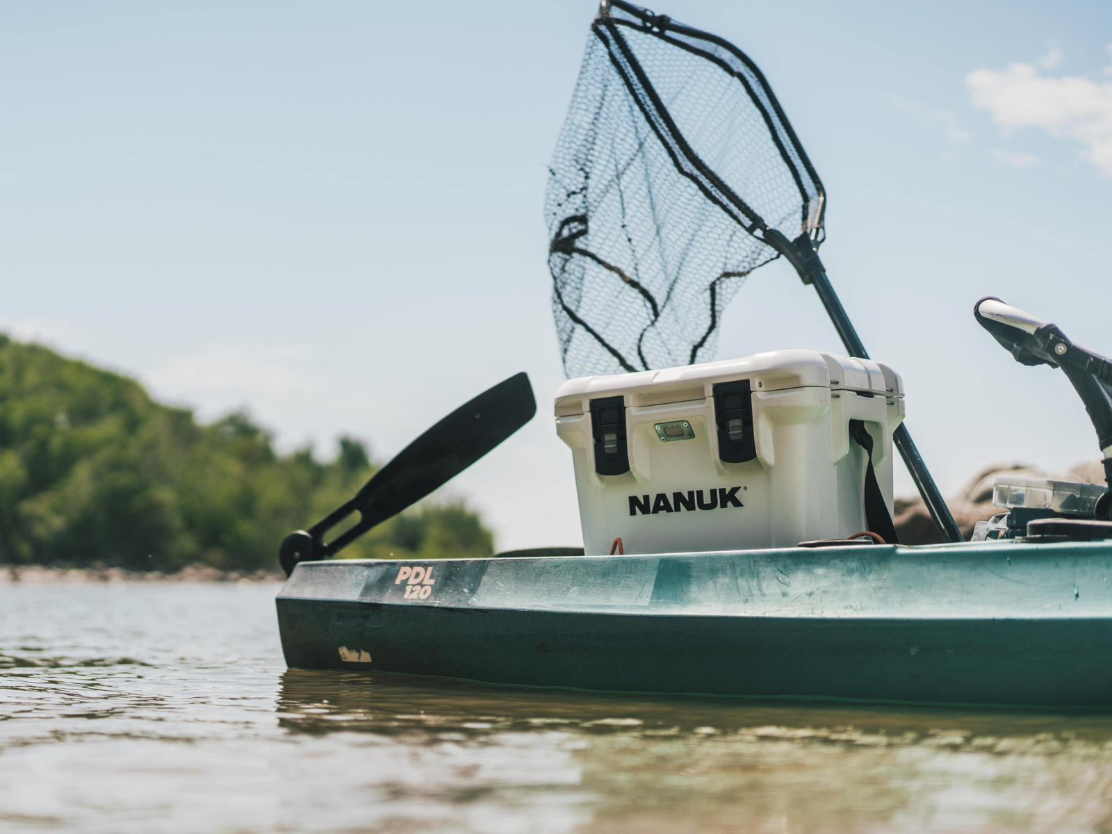 A white NANUK cooler box mounted on the deck of a green kayak on calm water, with a black landing net and paddle visible in the background.