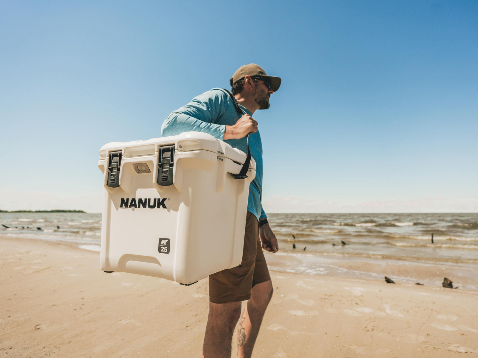 A man carrying a large white Nanuk cooler on his shoulder while walking along a sandy beach under a clear blue sky.