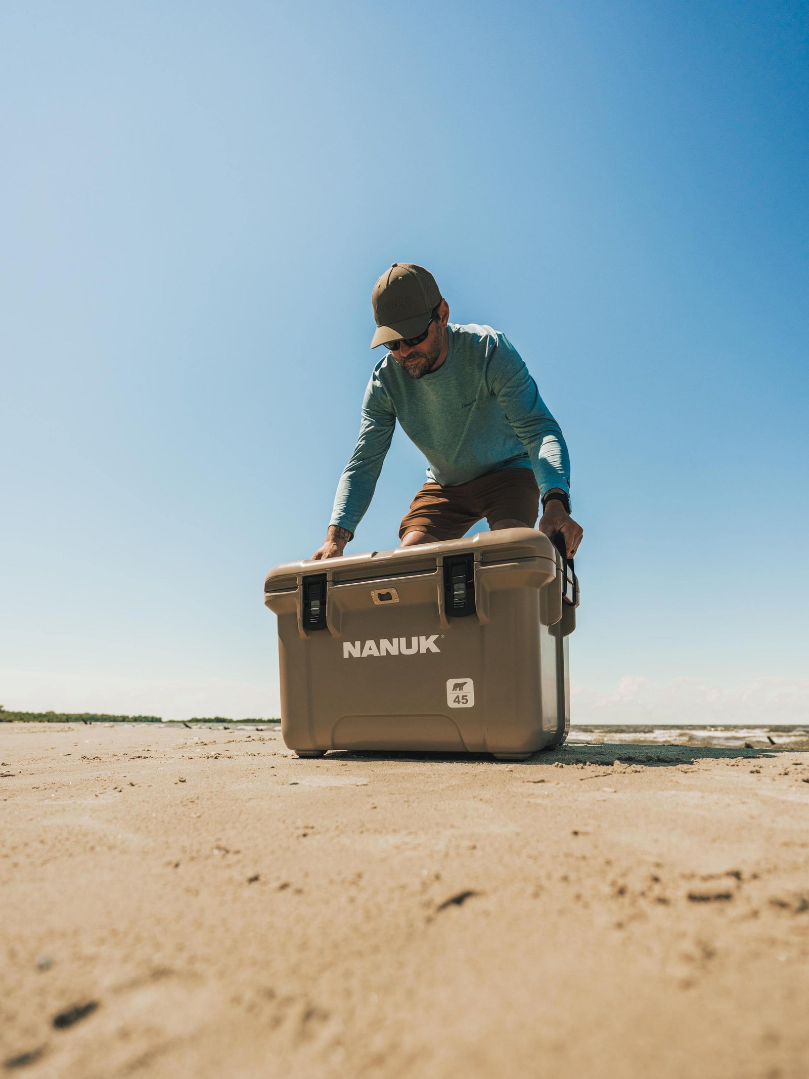 A man in a teal shirt and brown cap pushes a large beige Nanuk cooler on a sandy beach under a clear blue sky.
