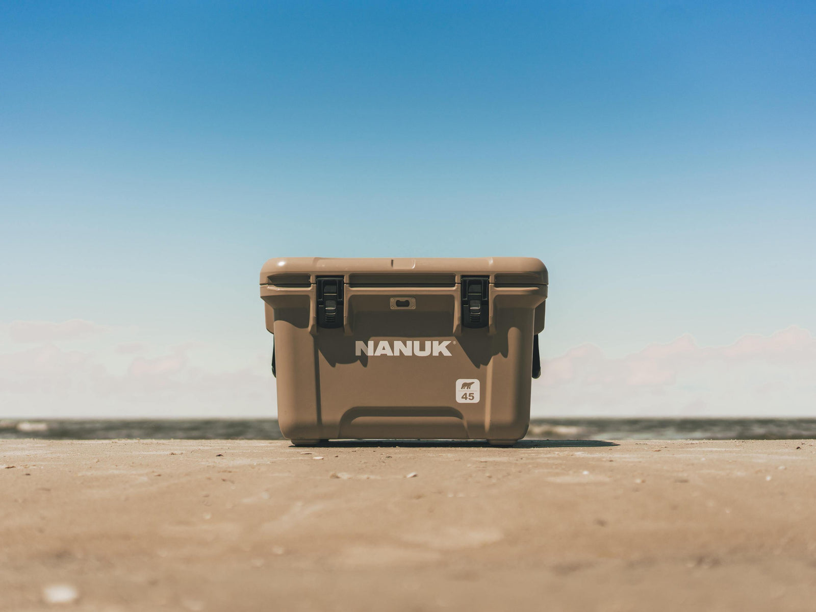 A tan Nanuk cooler box standing upright on a sandy beach with a clear blue sky in the background.