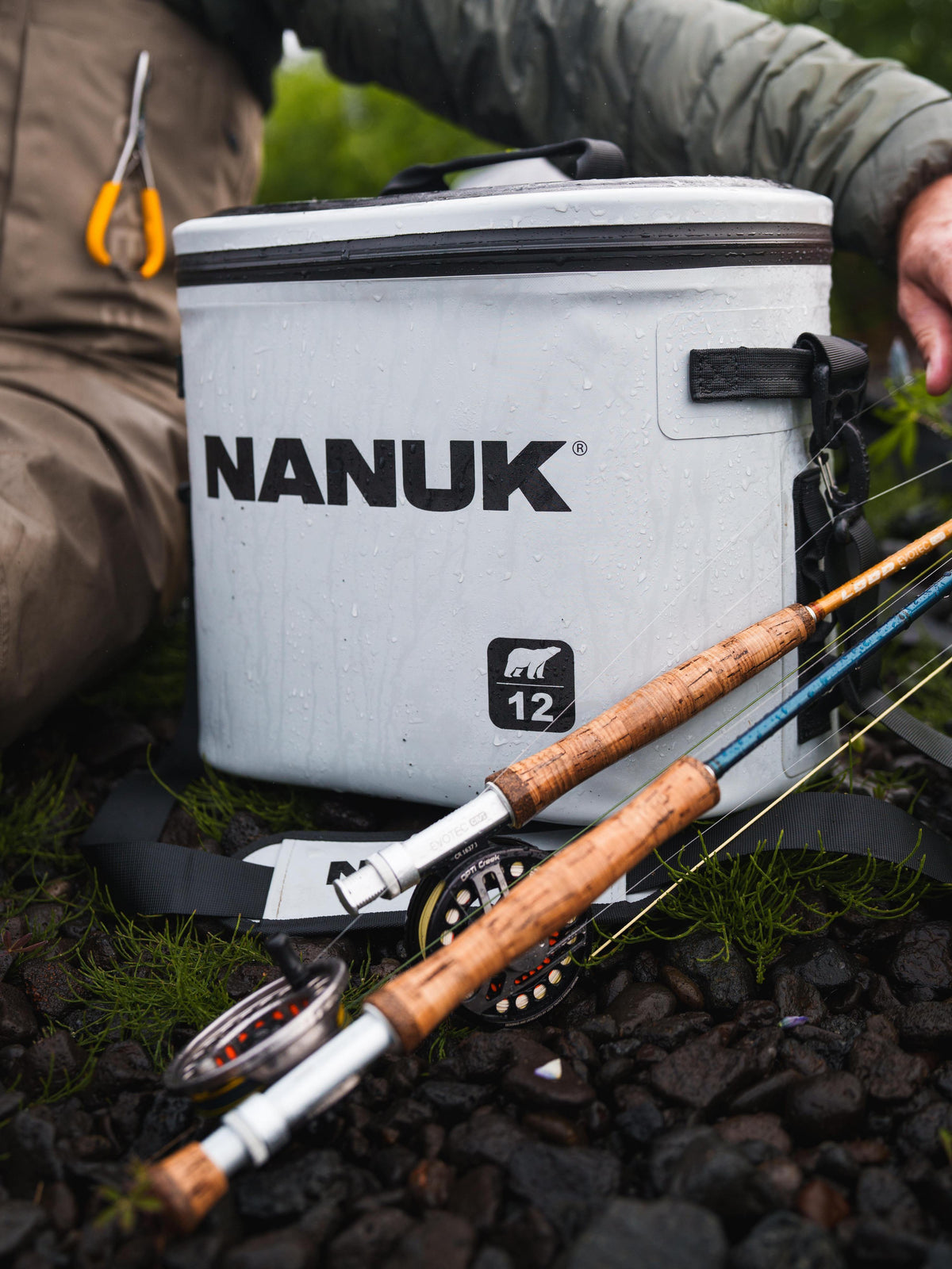 A white Nanuk 12-liter soft cooler bag sits on a rocky outdoor ground next to a person's legs and arms, with two fly fishing rods laid out in front of it.