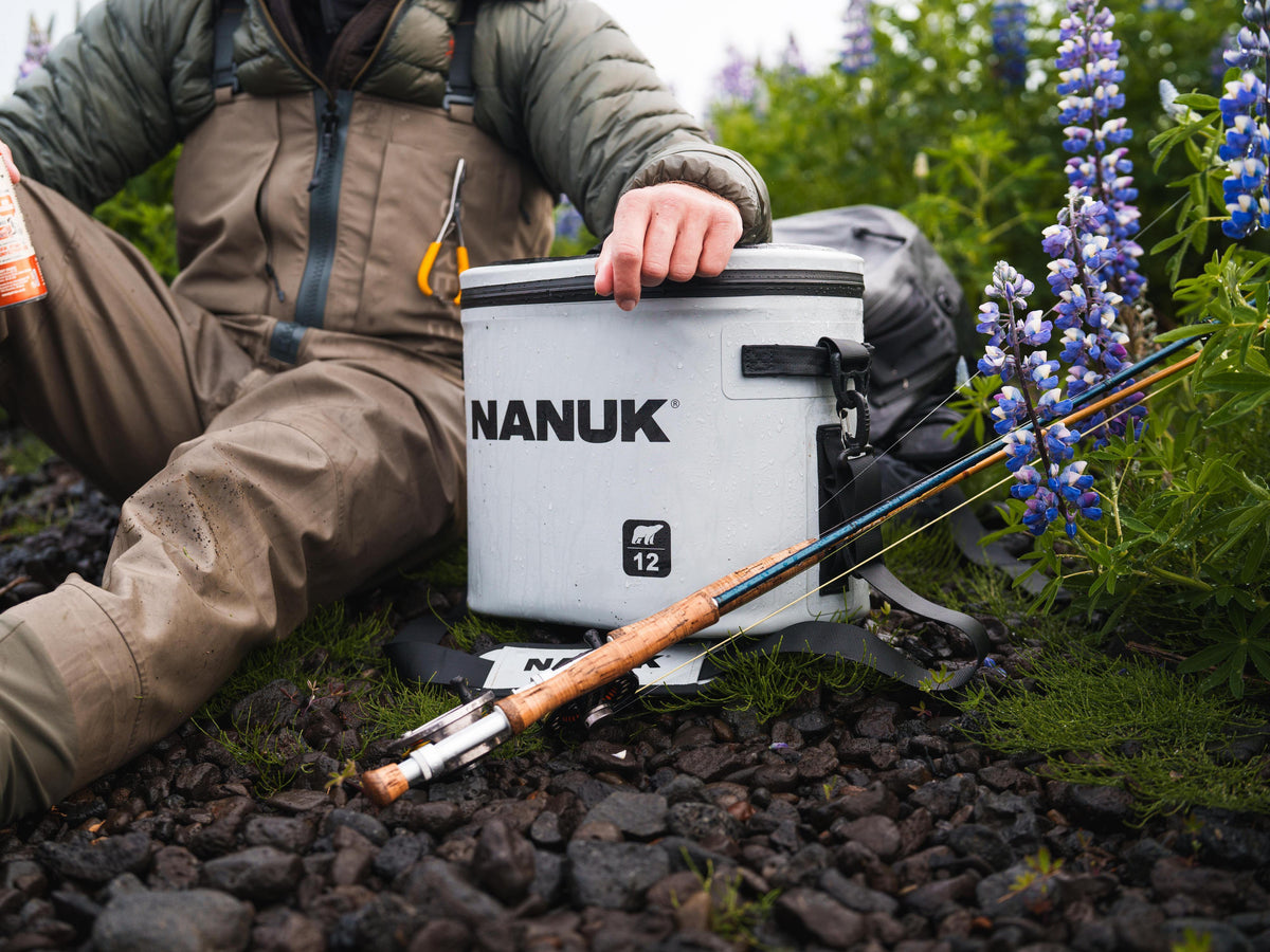 A person in fishing waders sits outdoors on dark gravel beside a white Nanuk cooler, with a fishing rod resting in front and blue-purple flowers in the background.