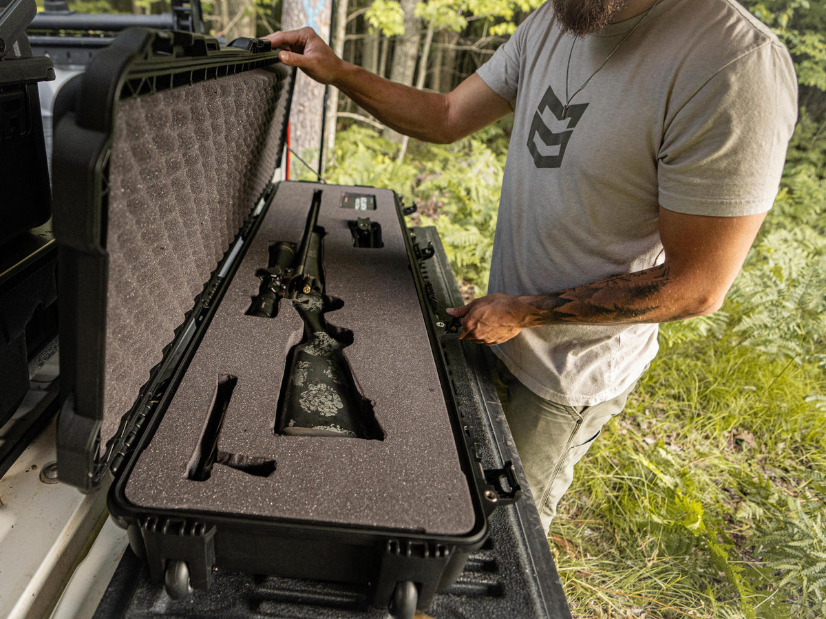 A man outdoors opens a black hard carrying case next to a vehicle, revealing a foam-lined compartment holding a rifle and accessories.