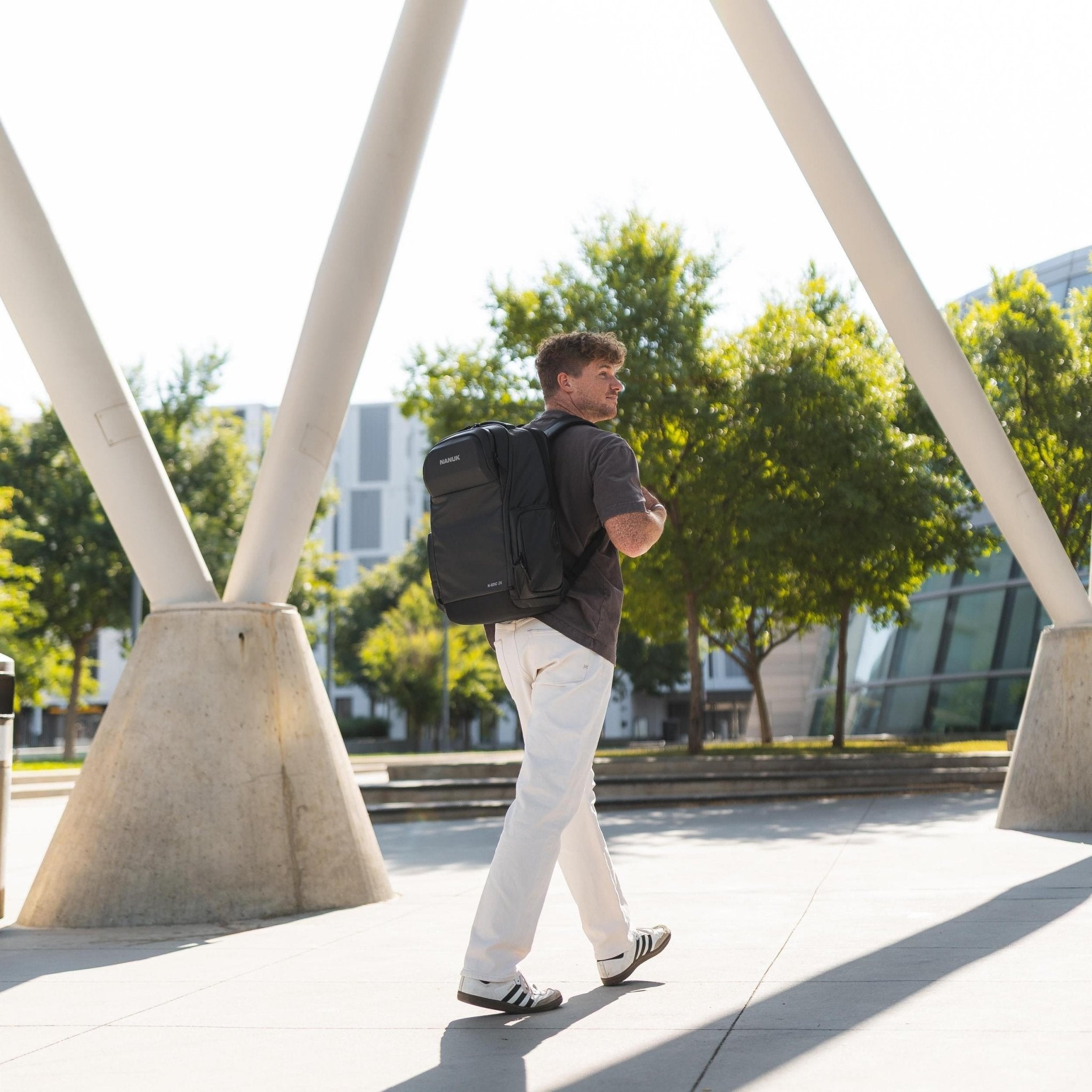 Man walking outdoors with a backpack, surrounded by trees and modern architecture. Man walking outdoors with a backpack, surrounded by trees and modern architecture.