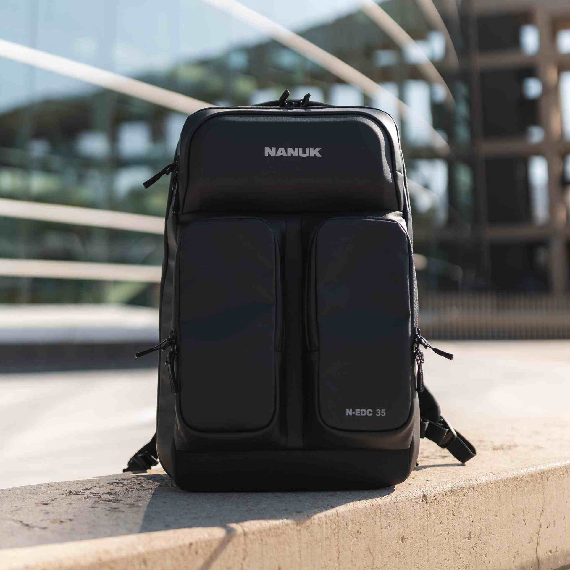 Black backpack with 'NANUK' branding on a concrete ledge with a modern building in the background. Black backpack with 'NANUK' branding on a concrete ledge with a modern building in the background.