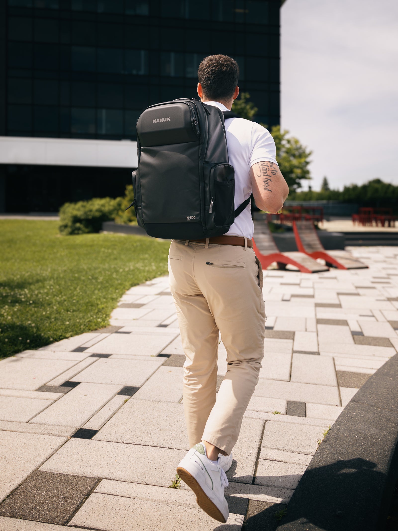Man walking outdoors with a black backpack, wearing a white shirt and beige pants.