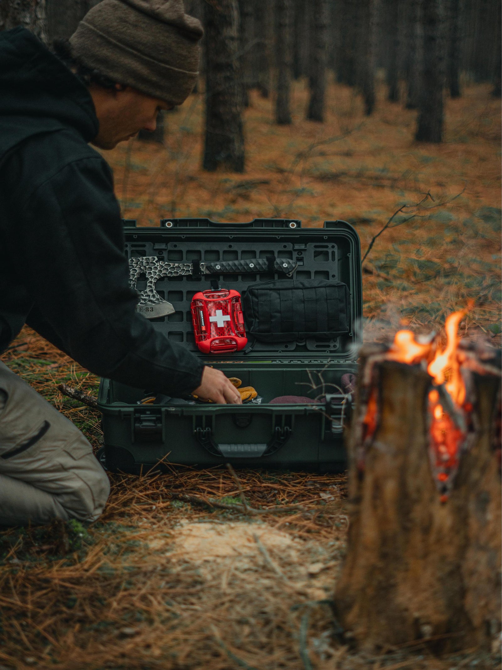Person in a forest setting with a black toolbox and red first aid kit near a campfire.