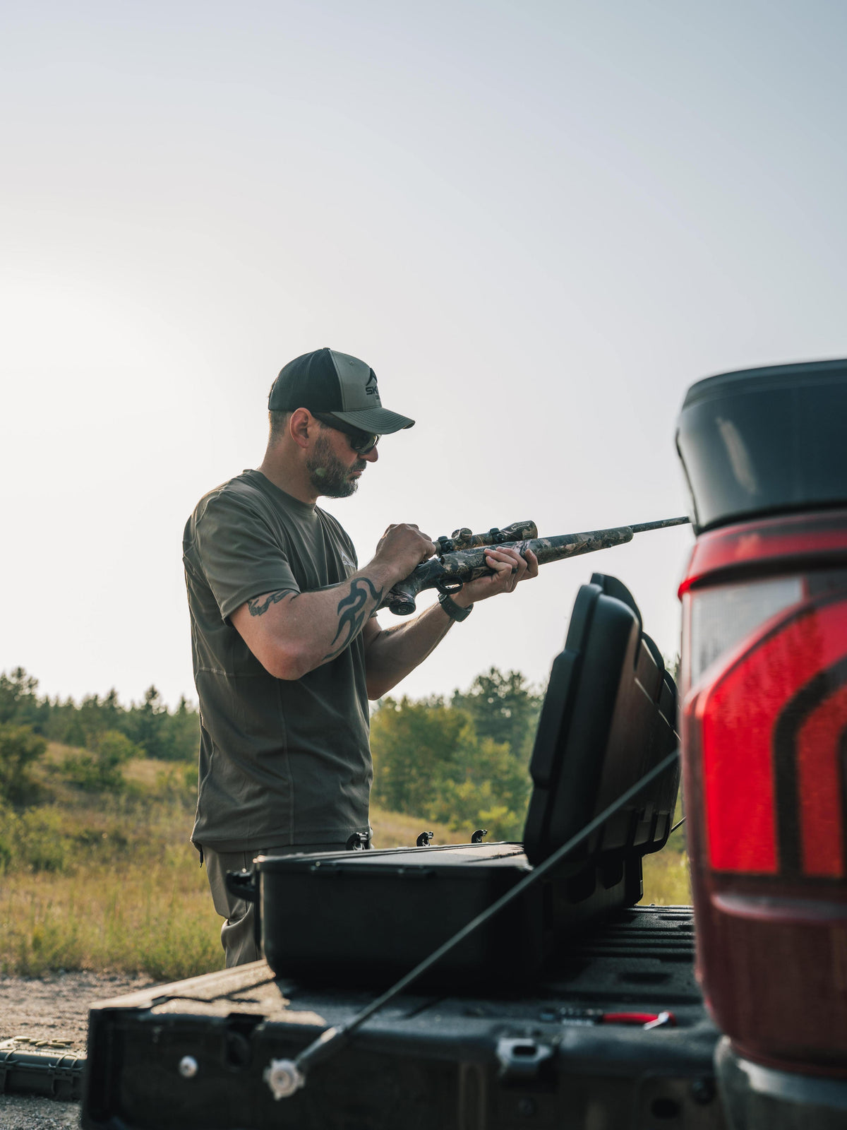 A man wearing a dark cap and t-shirt stands beside an open black hard case on the tailgate of a red truck, holding a rifle in an outdoor field setting.