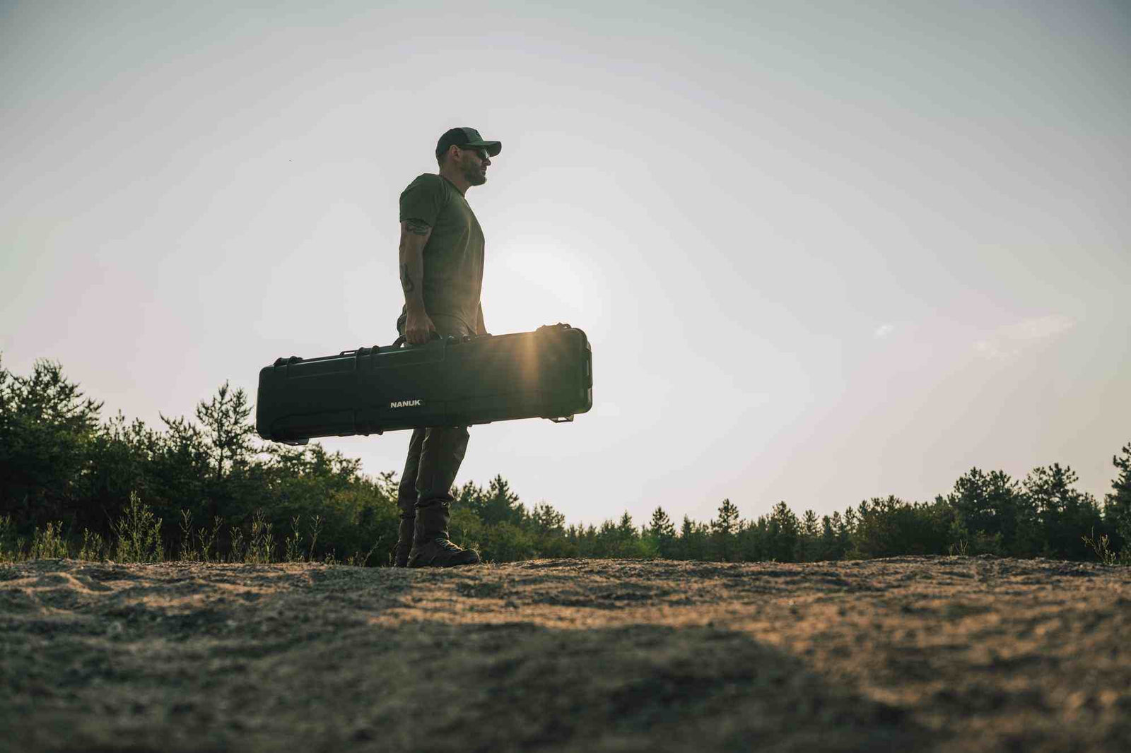 Person holding a large black case against a natural background with trees and open sky.