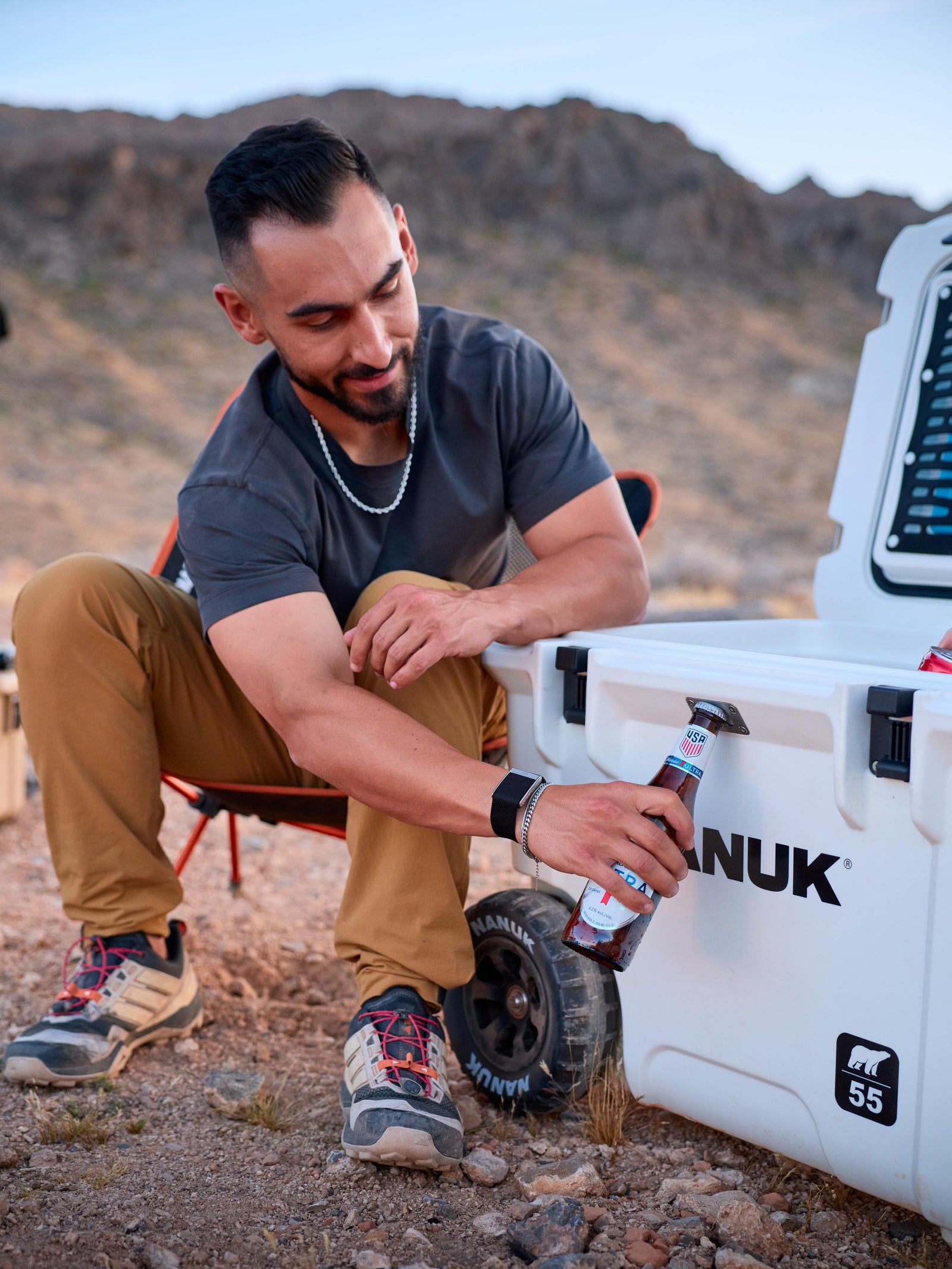 Man sitting by a white vehicle in a desert setting, holding a can of Anuk.