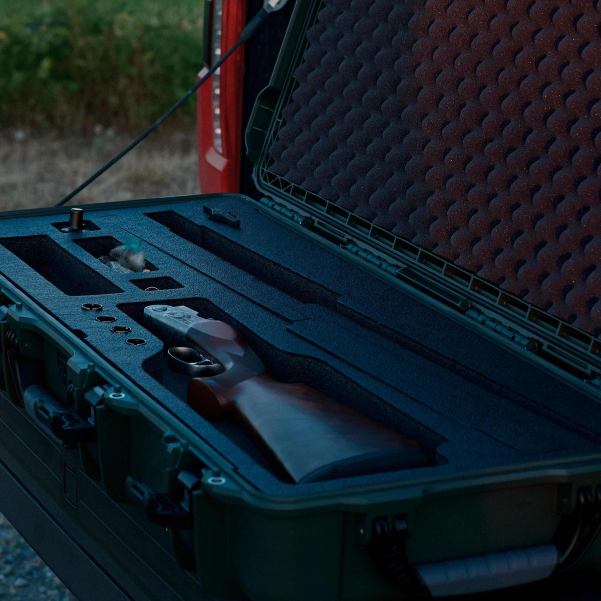 A shotgun resting inside a black foam-lined hard carrying case, shown open outdoors with a blurred red vehicle and foliage in the background.