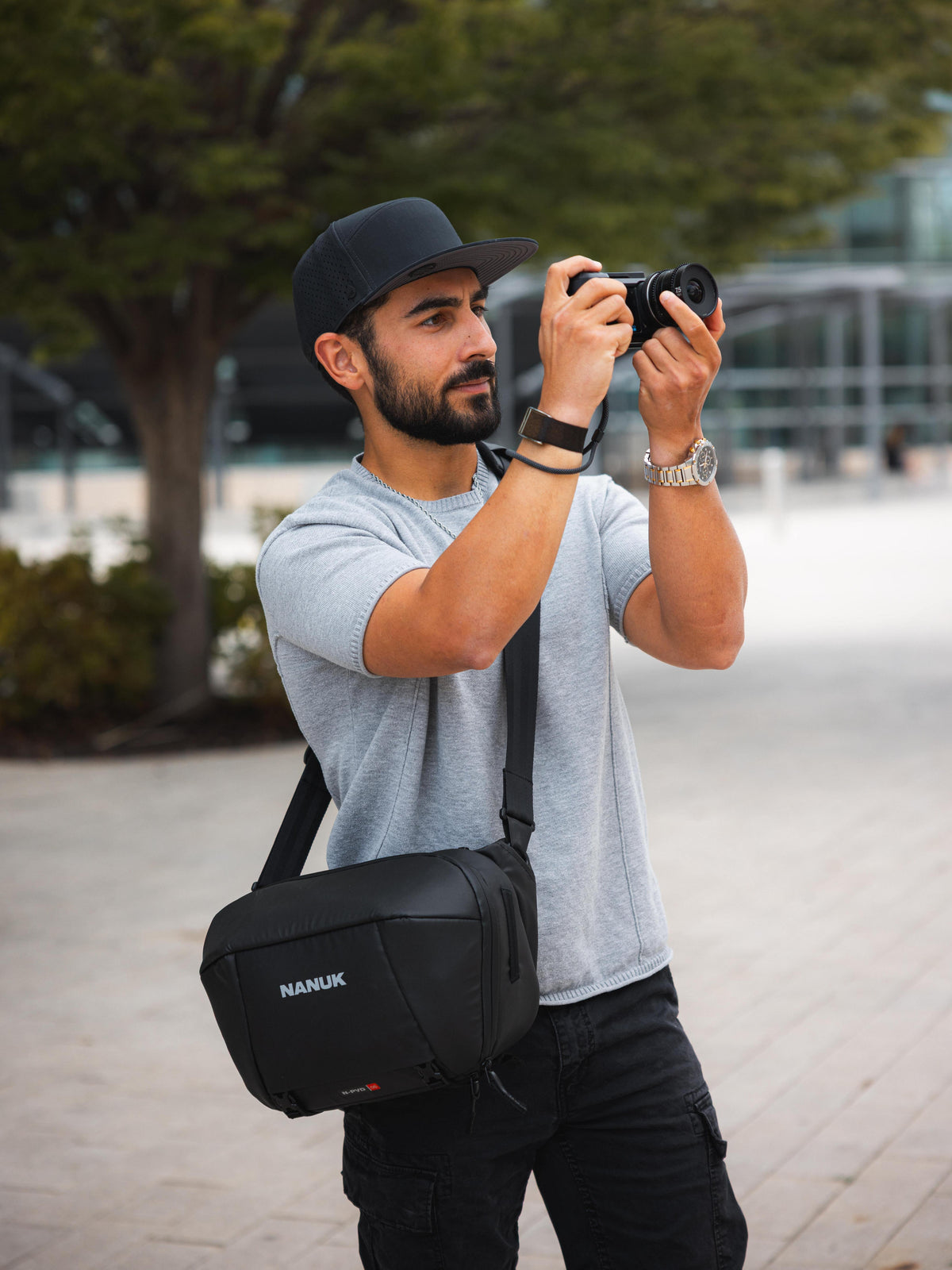 A man wearing a gray t-shirt, black cap, and black cargo pants is holding a camera and wearing a black NANUK crossbody bag with a long strap, set against a blurred outdoor background.