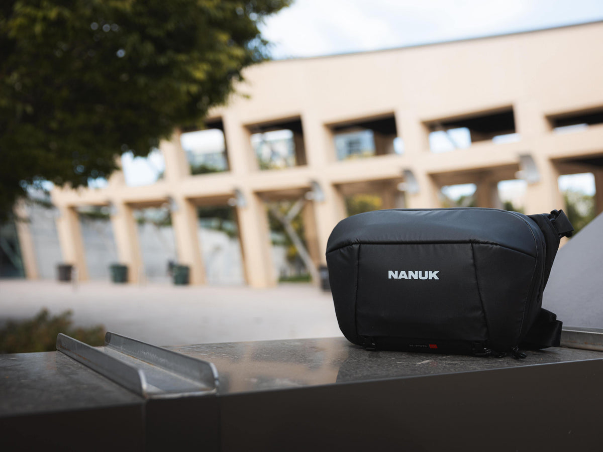 A black Nanuk camera bag with a white logo, resting on a dark metal surface in an outdoor setting with a blurred modern building in the background.