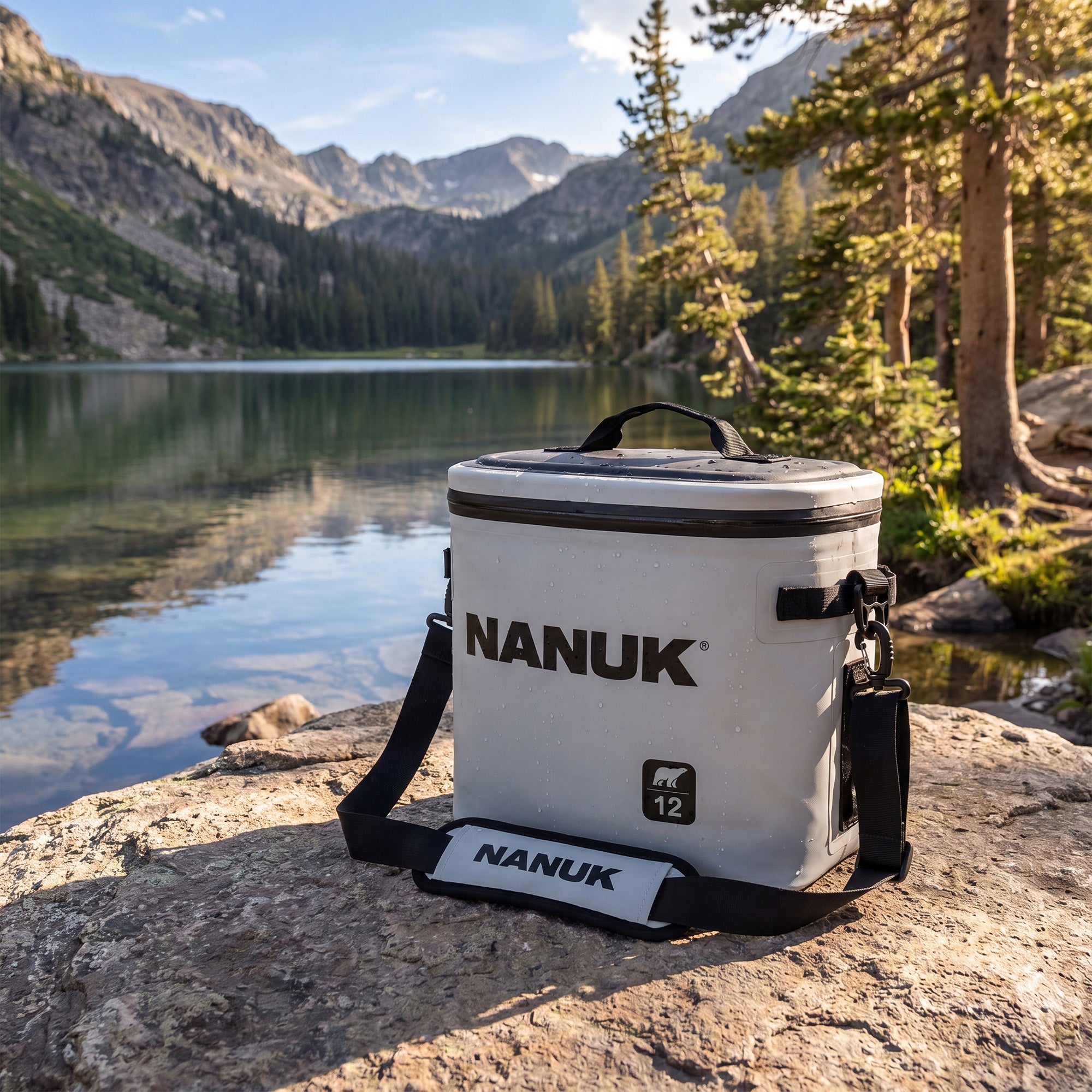 A white and black Nanuk 12-inch cooler bag sits on a large rock in a mountain lake setting, with pine trees and rugged peaks in the background. A white and black Nanuk 12-inch cooler bag sits on a large rock in a mountain lake setting, with pine trees and rugged peaks in the background.