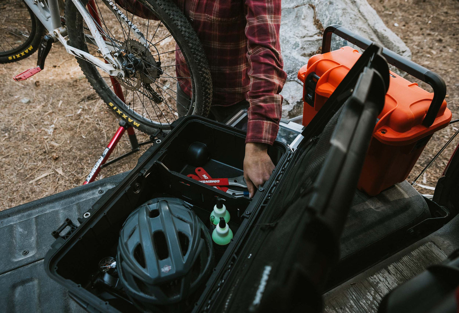 Person loading a black NANUK Trail 30 case into the bed of a pickup truck next to a mountain bike on a forest trail.