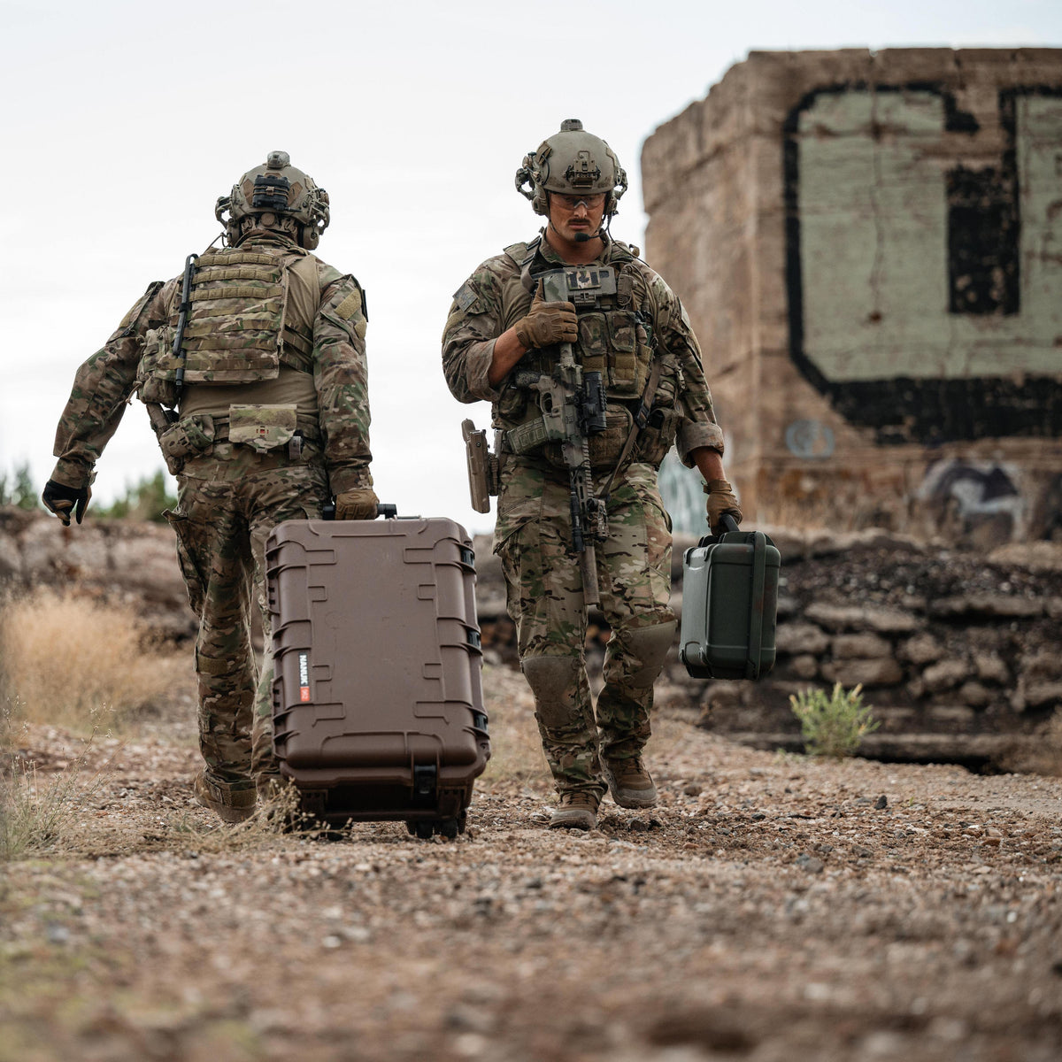 Two soldiers in camouflage uniforms walk through a dusty outdoor area, one pulling a large brown hard-sided case and the other holding a rifle and a small green case. A weathered building stands in the background.