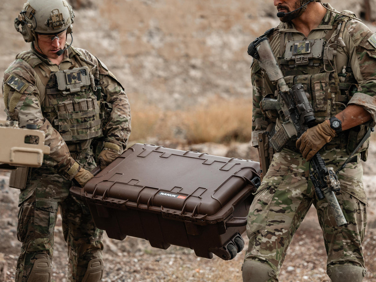Two soldiers in camouflage uniforms carry a large brown hard case with wheels across a rocky outdoor terrain. One soldier holds the left side of the case, and the other stands beside it holding a rifle.