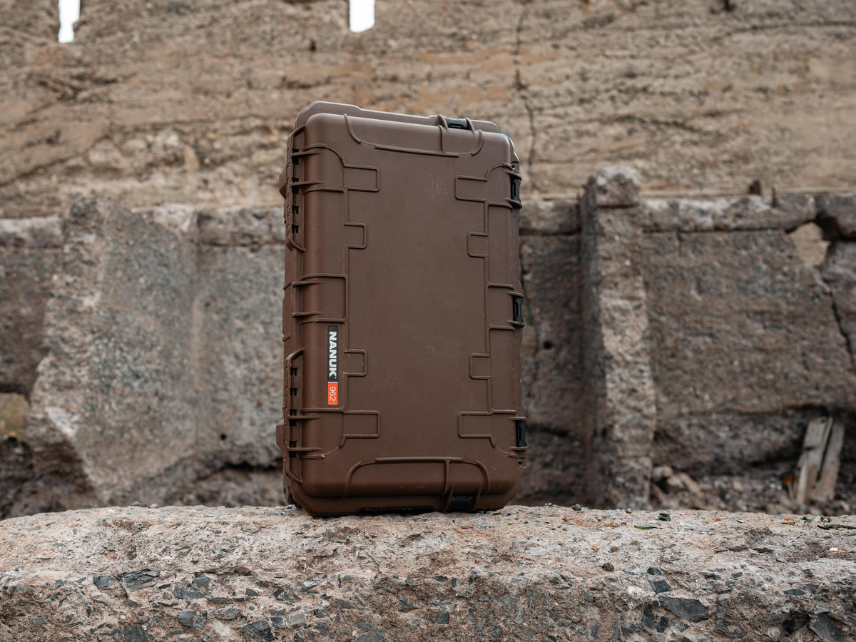 A brown hard protective case standing upright on a stone surface against a rustic stone wall background. The case has visible hinge latches on the sides and a small red/orange vertical logo label that reads NANUK