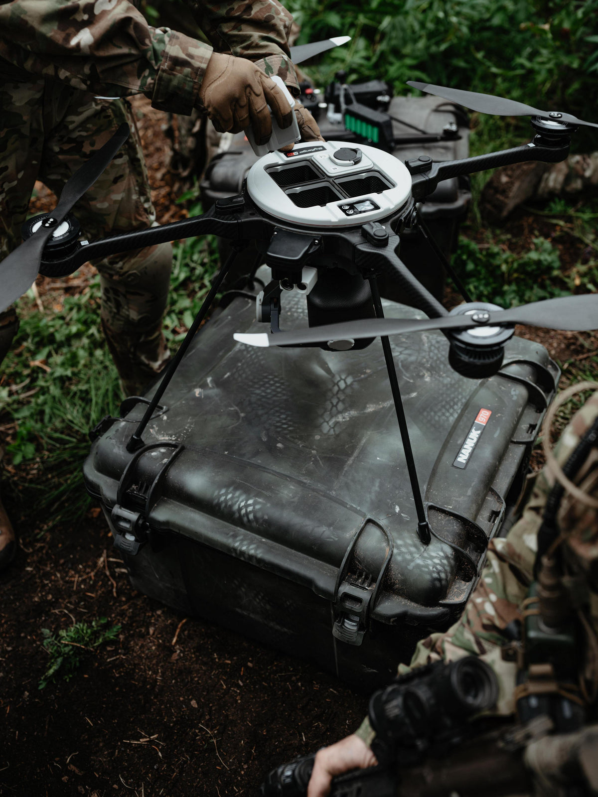 A black rugged hard case secured to a quadcopter drone in an outdoor forest setting, with people in camouflage clothing handling the setup.