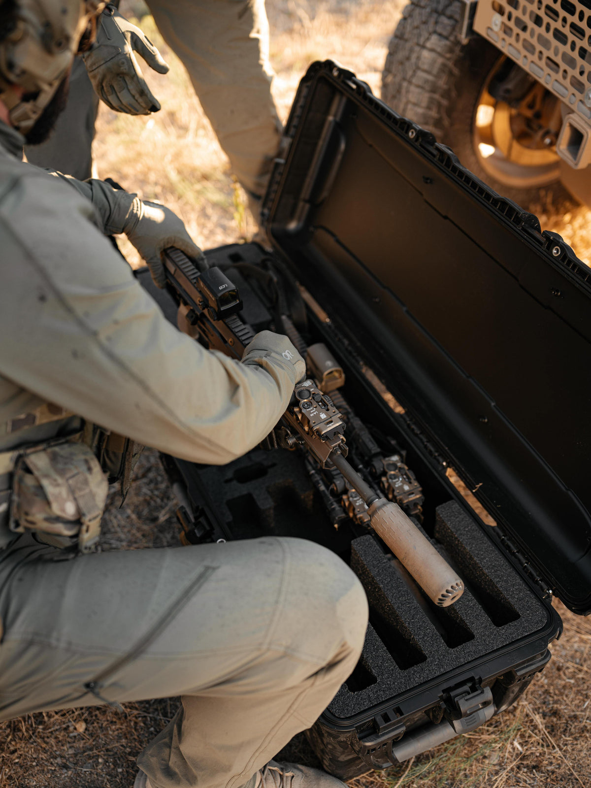 A black hard carrying case opened on a dry outdoor ground, containing a rifle with a mounted scope and a sand-colored silencer. A person in tan tactical clothing is kneeling beside the case, and another person is partially visible at the top near a vehicle tire.
