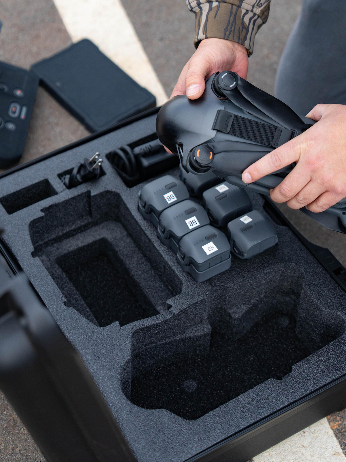 A close-up of a person holding a black drone controller above a custom foam storage tray filled with multiple battery packs inside an open hard case.