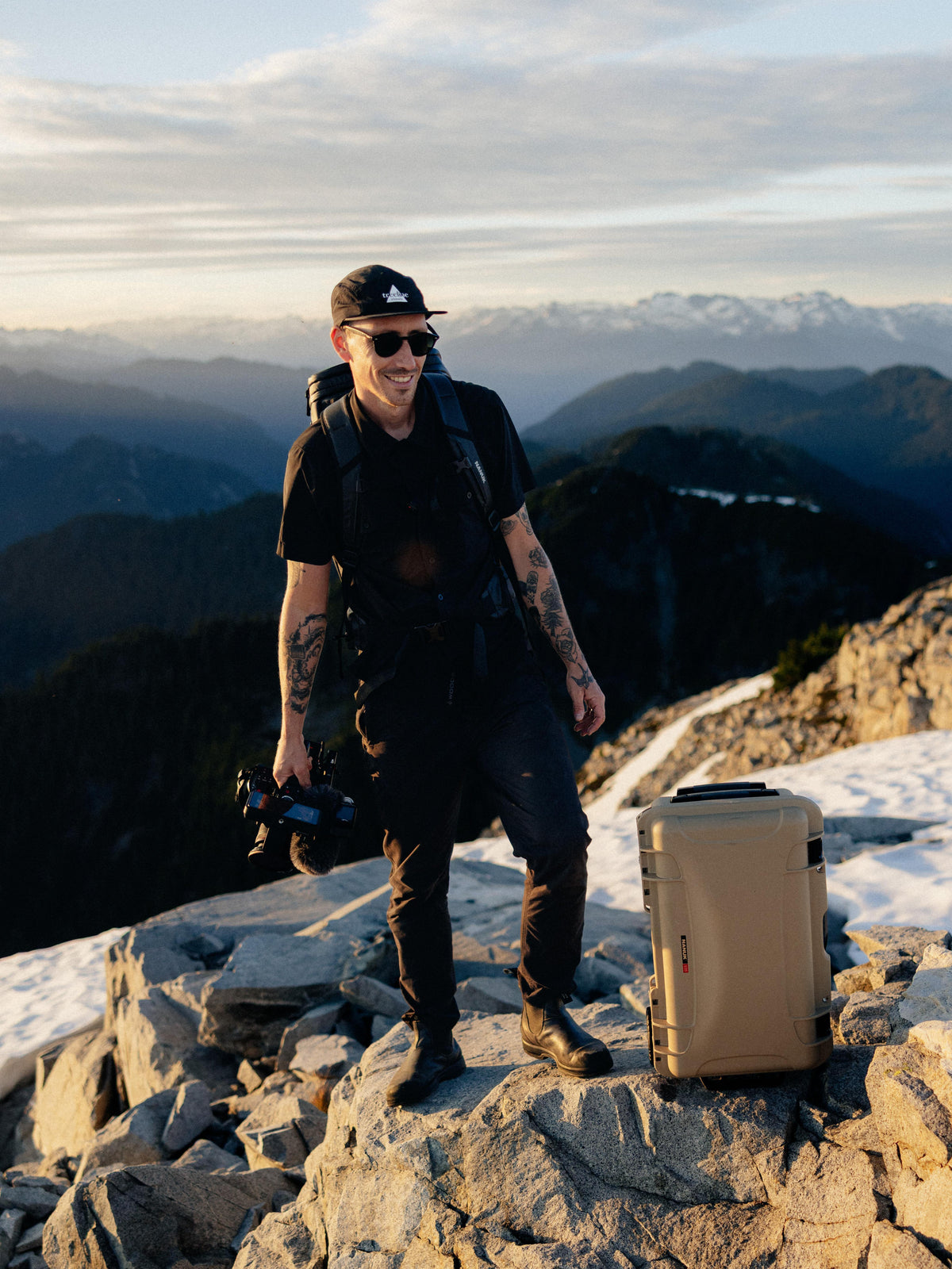 A man stands on a rocky mountain peak with a scenic mountain range and sky in the background. He is wearing a black cap, sunglasses, black t-shirt, and black overalls, holding a camera, with a beige hard-sided case standing next to him.