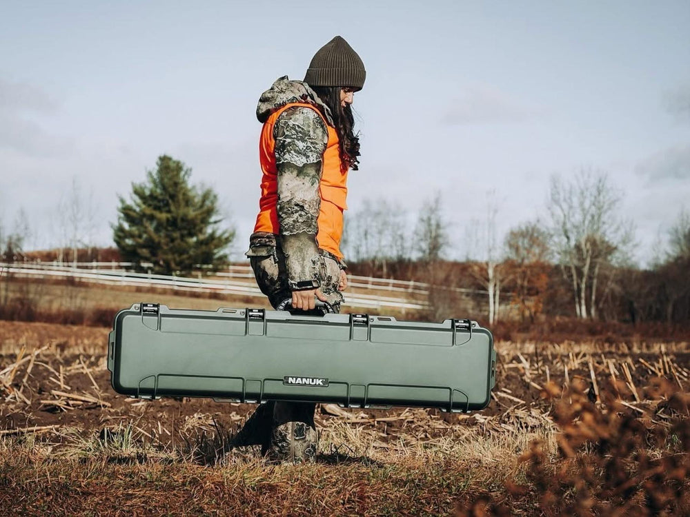 Person in hunting gear holding a large green hard-shell case in a field.