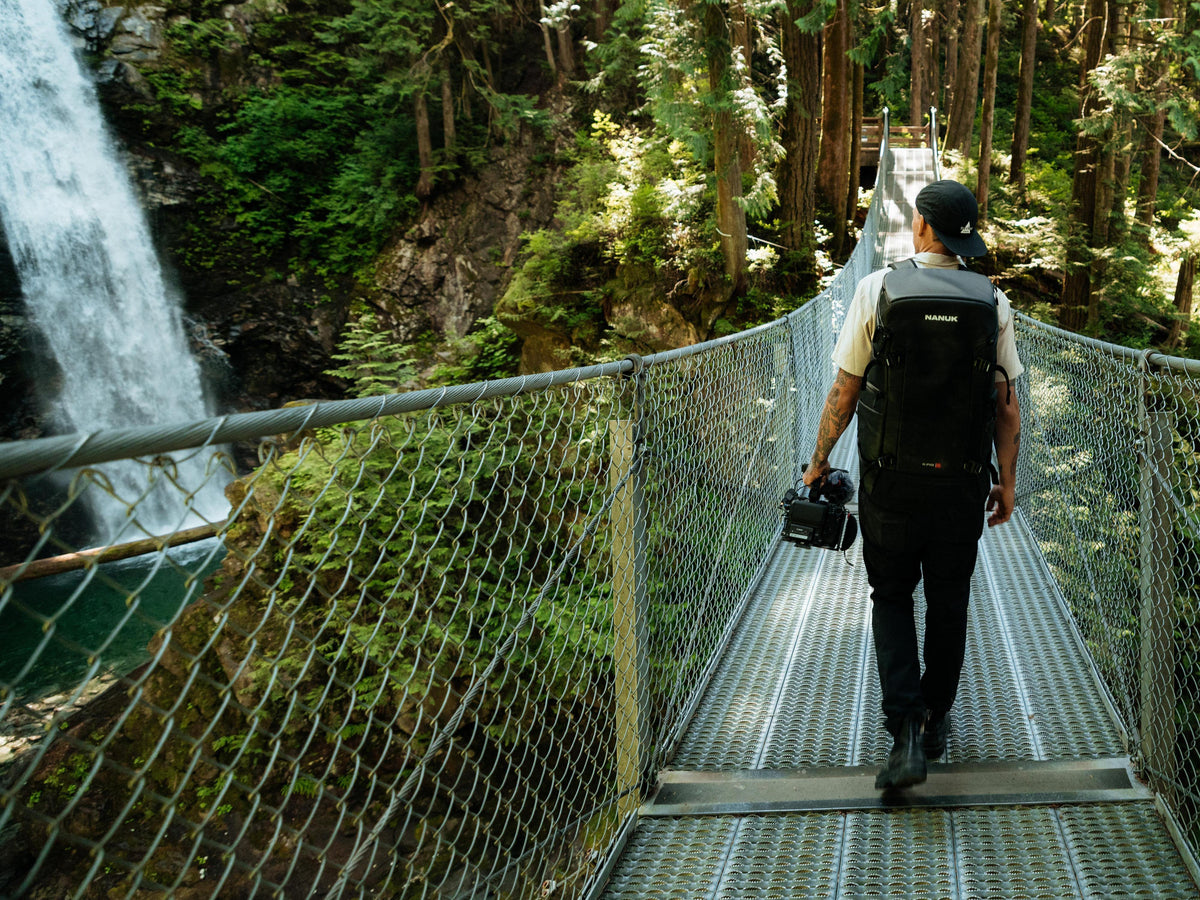A man wearing a black backpack and walking across a suspension bridge in a forest, with a waterfall visible on the left side. He is carrying a camera.