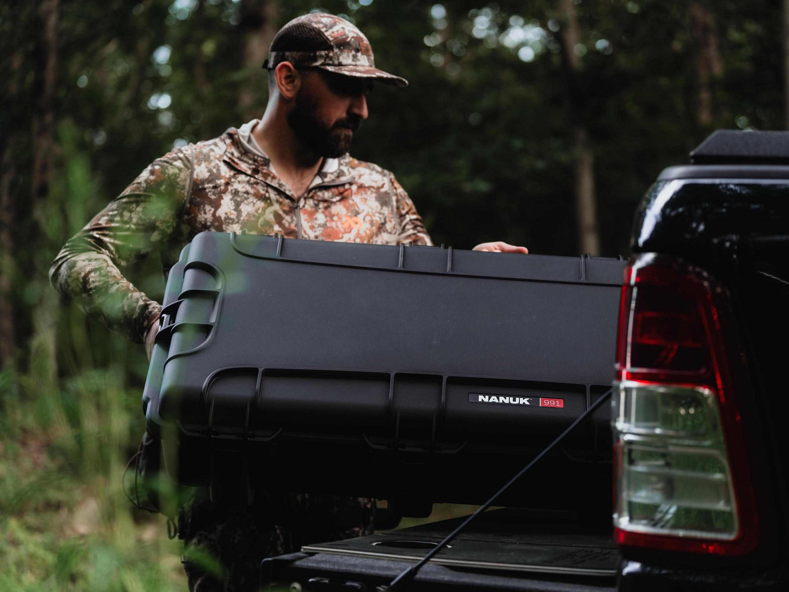 Man in camouflage holding a large black Nanuk toolbox next to a pickup truck in a forest setting