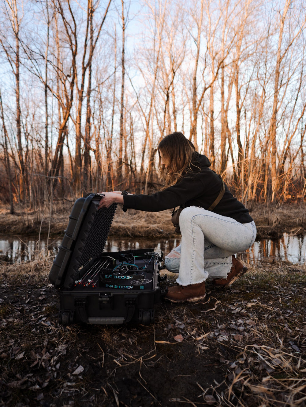 Person sitting by a pond with a fishing rod and tackle box in a forest setting