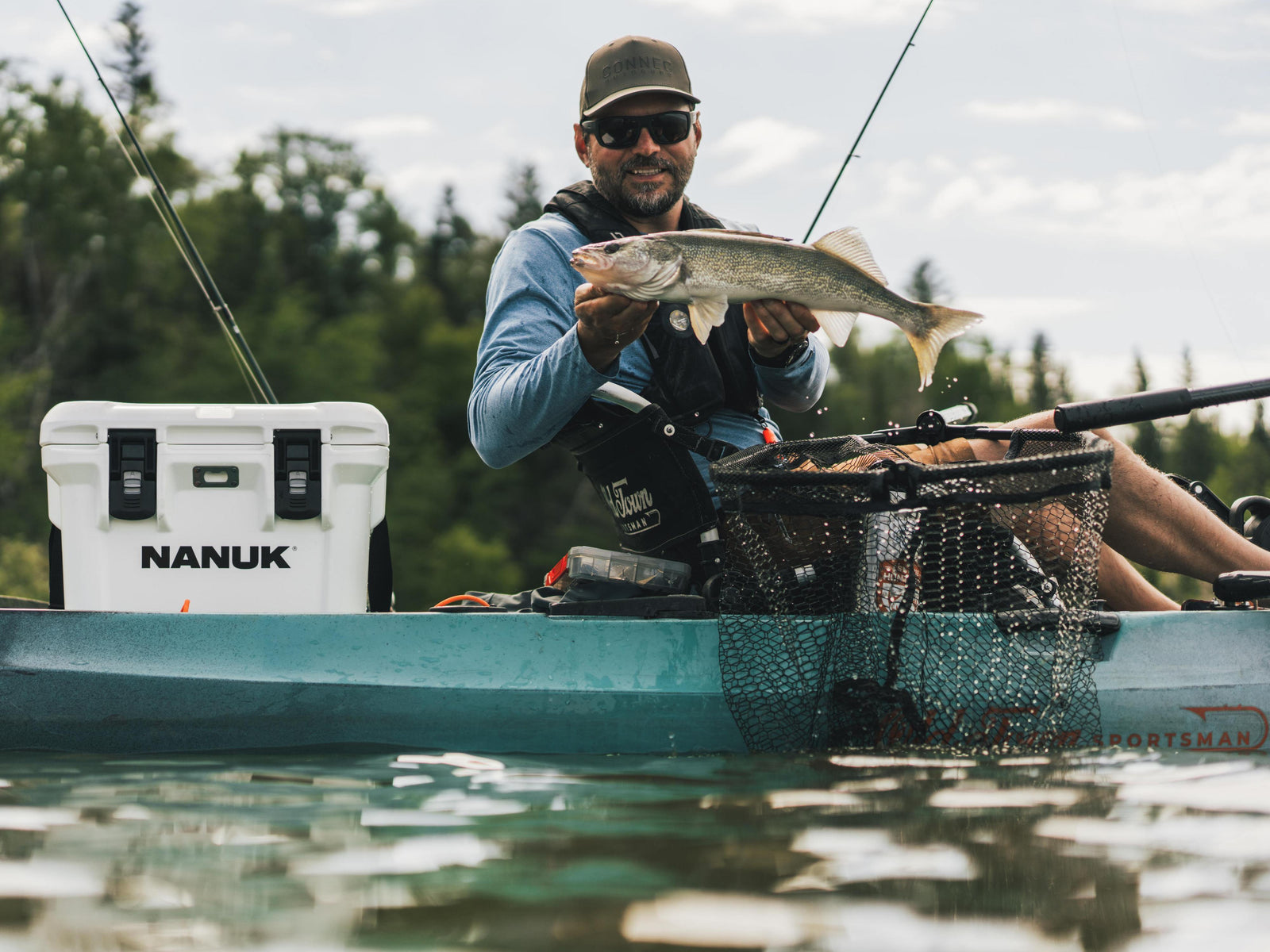 A man in a teal kayak on the water holds a large fish while wearing a black Nanuk life vest. A white Nanuk cooler is sitting to his left, and a black landing net is in front of him.
