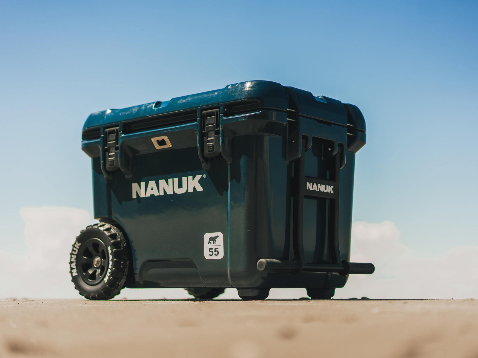 A dark teal Nanuk cooler with rugged off-road wheels, shown outdoors on a sandy surface under a clear blue sky. The cooler features the brand name 'NANUK' in white on the side and a small '55' label.