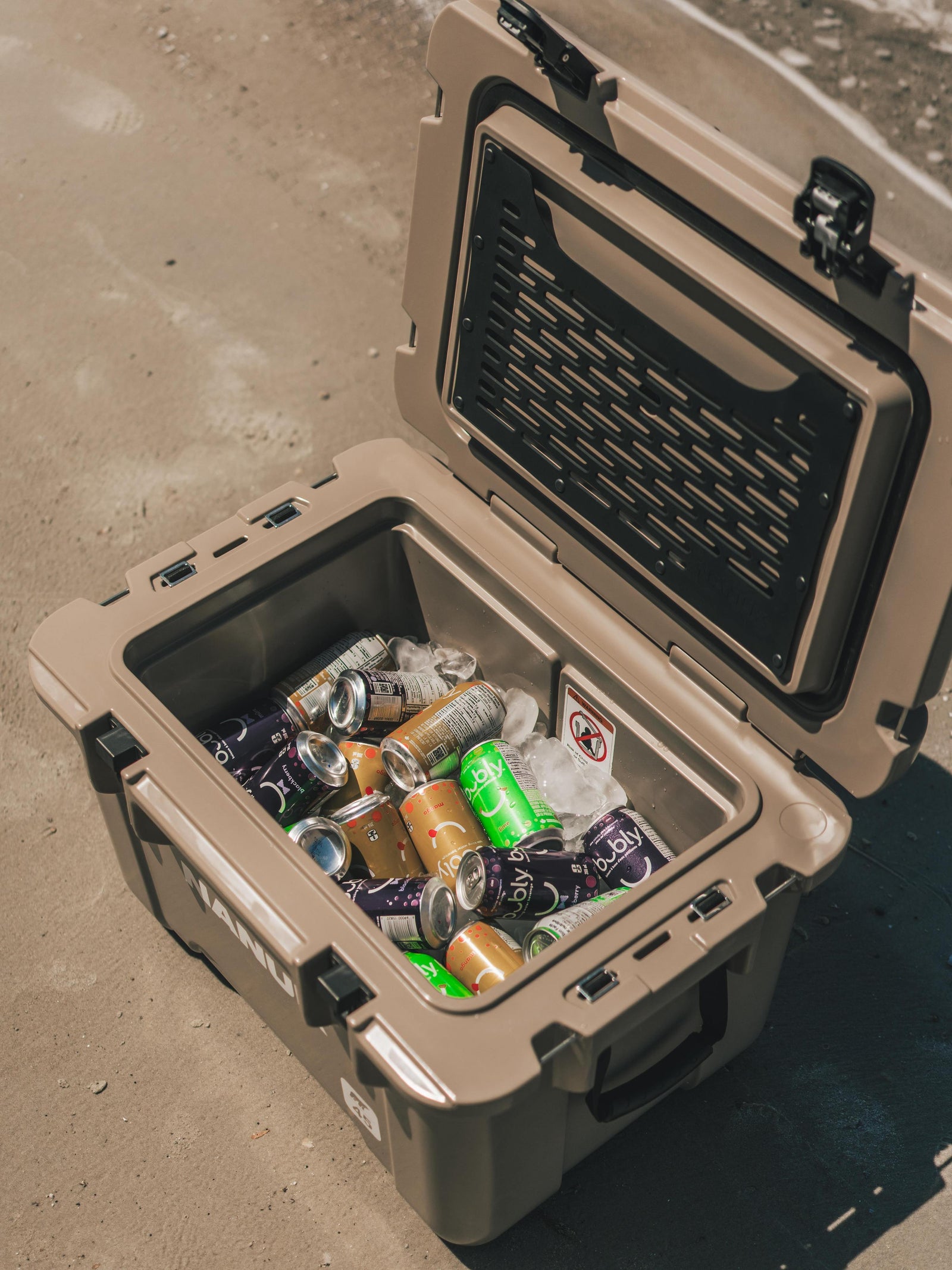 A tan hard-sided cooler with its lid open, filled with crushed aluminum cans and ice, placed on a concrete surface.