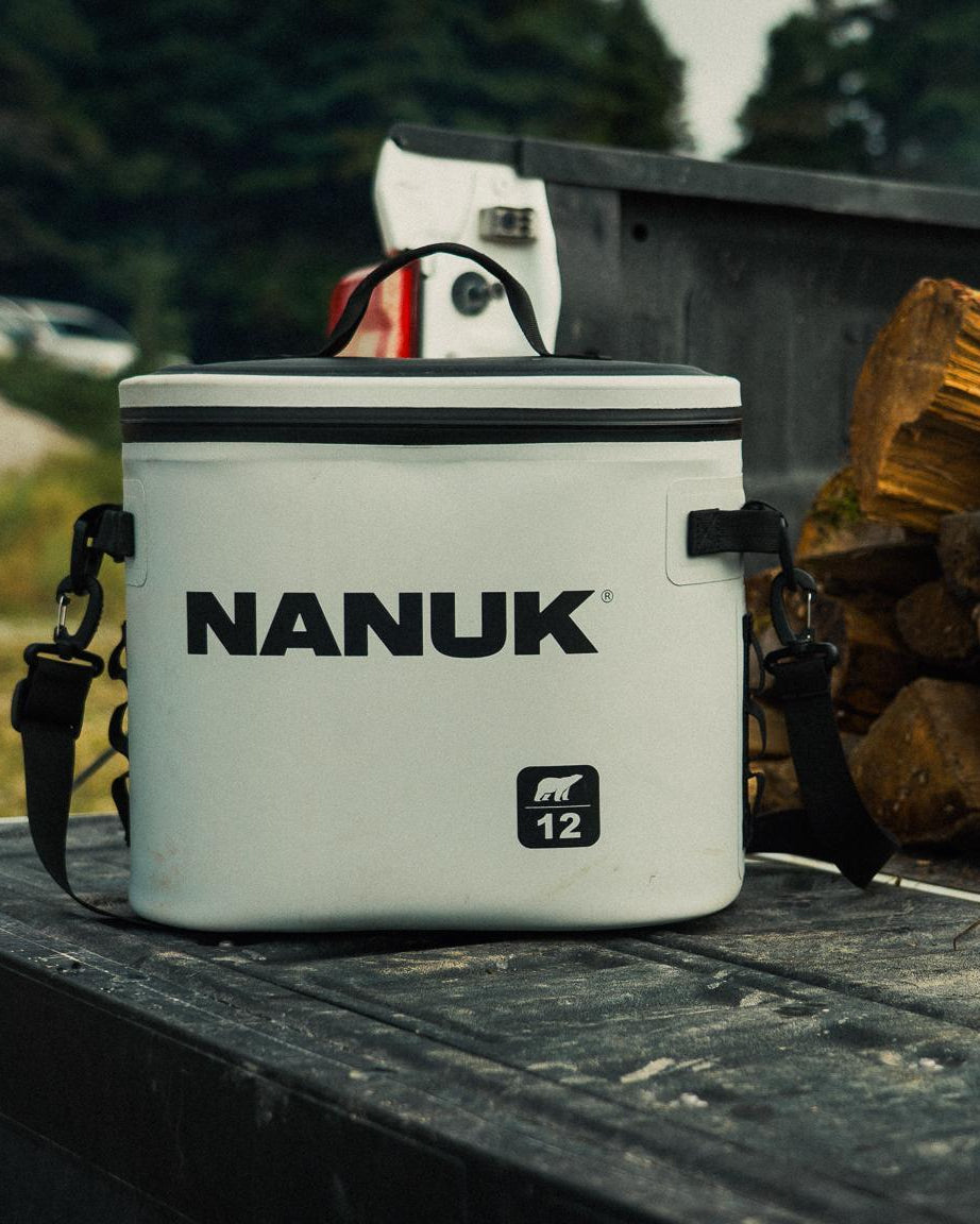 A white Nanuk 12-liter cooler with black straps and branding sits on the bed of a truck next to a stack of firewood.