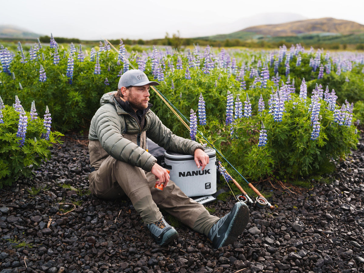 A man sits on dark gravel in a field of purple lupines, holding fishing rods next to a white Nanuk cooler. He is wearing a gray puffer jacket, khaki pants, and a gray cap.
