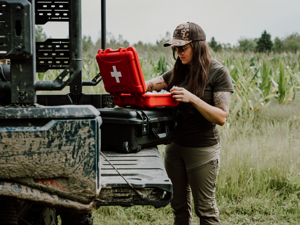 A woman in outdoor work clothes stands beside a utility vehicle tailgate in a field, handling a red first-aid case that is resting on top of a black hard case.