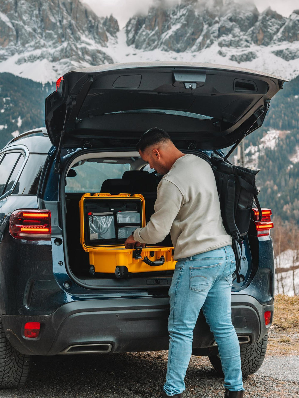 Person loading a yellow suitcase into the trunk of a car with mountains in the background