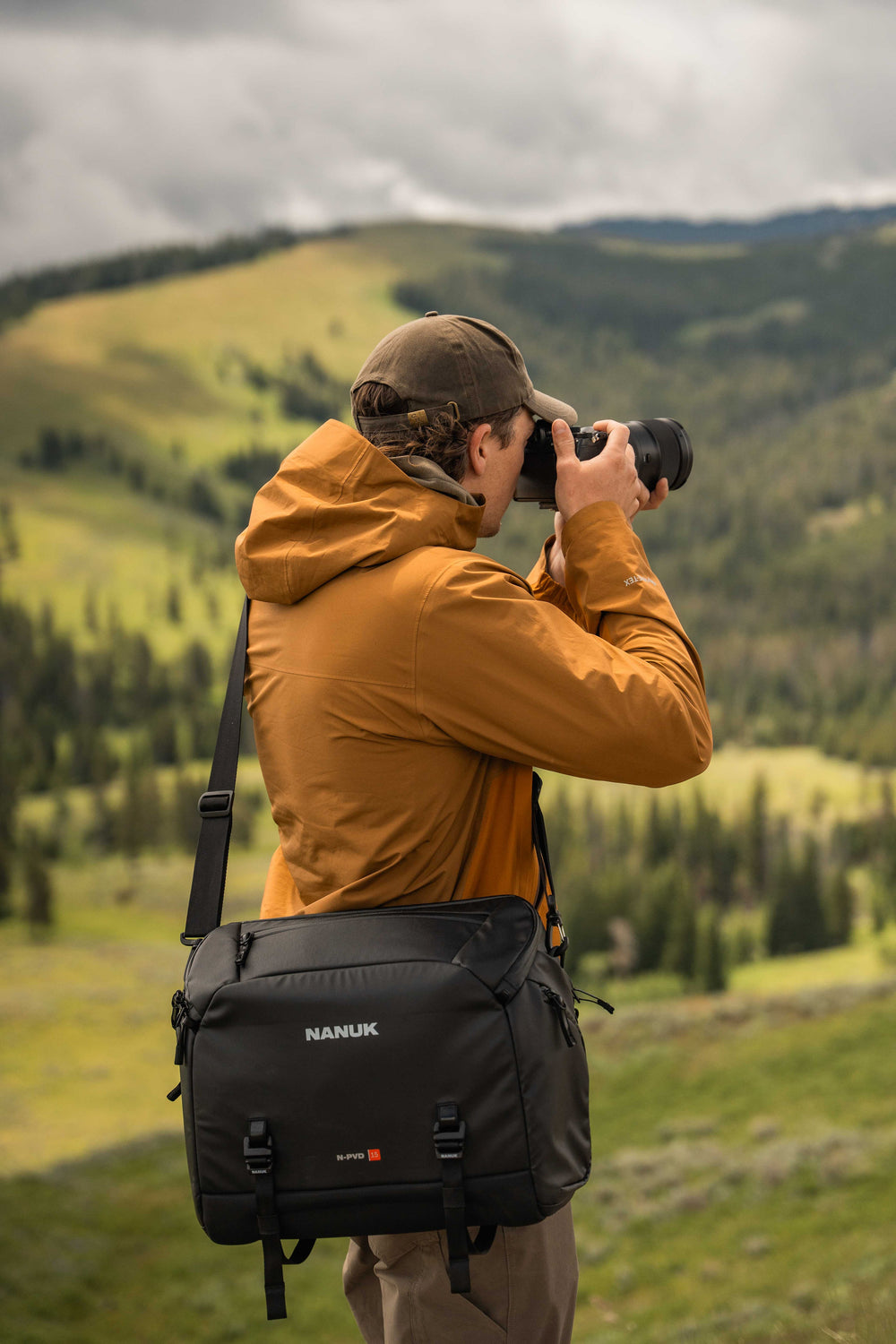 Person taking a photo with a camera in a mountainous landscape, wearing an orange jacket and carrying a black Manfrotto bag.