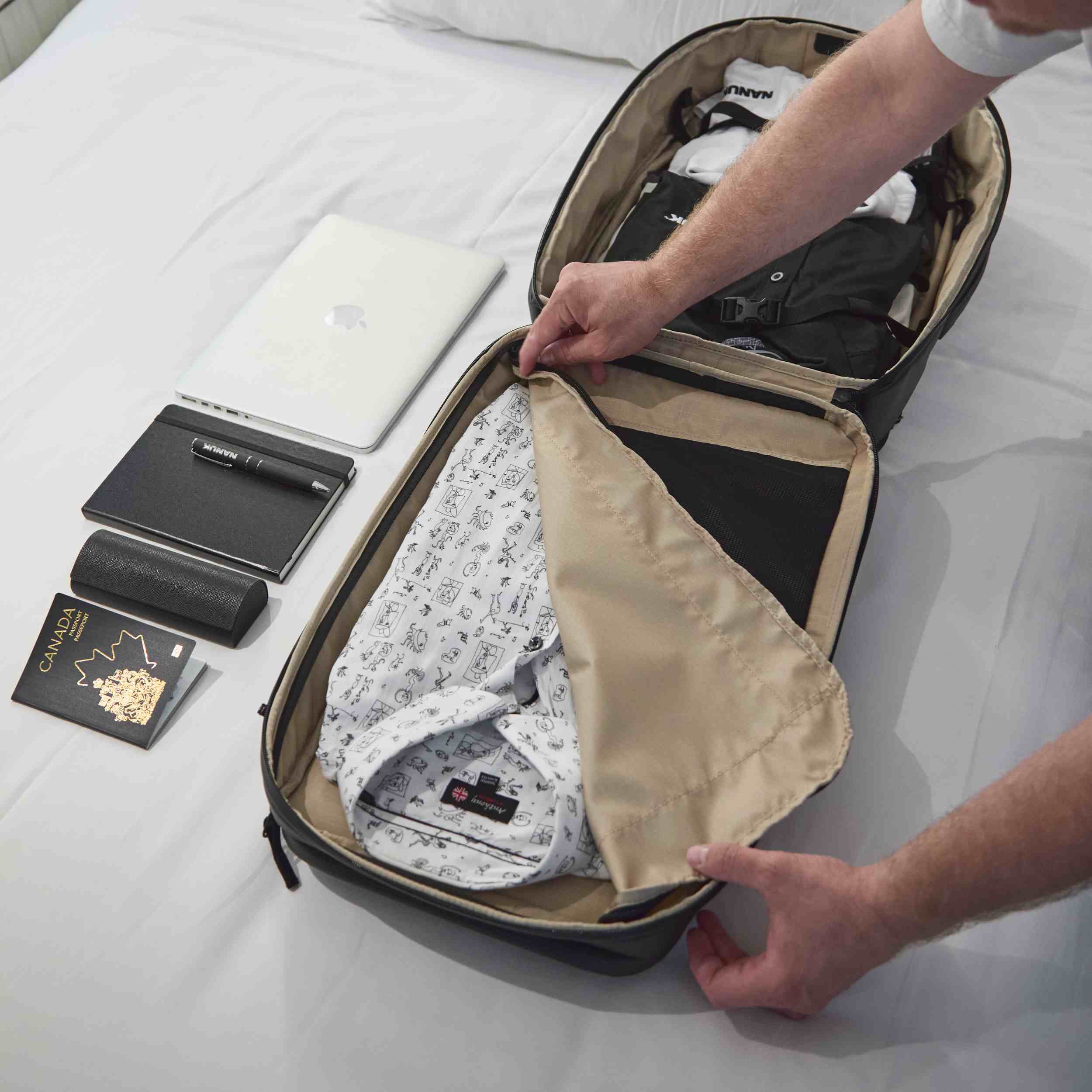 Person packing clothes into a suitcase on a bed with a laptop and other items nearby.