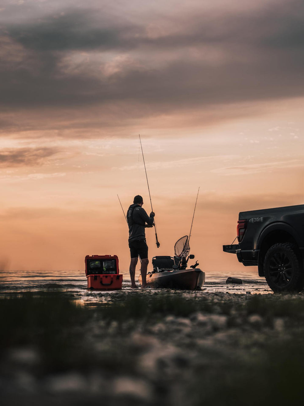 Person fishing from a kayak on a beach at sunset with a truck nearby.