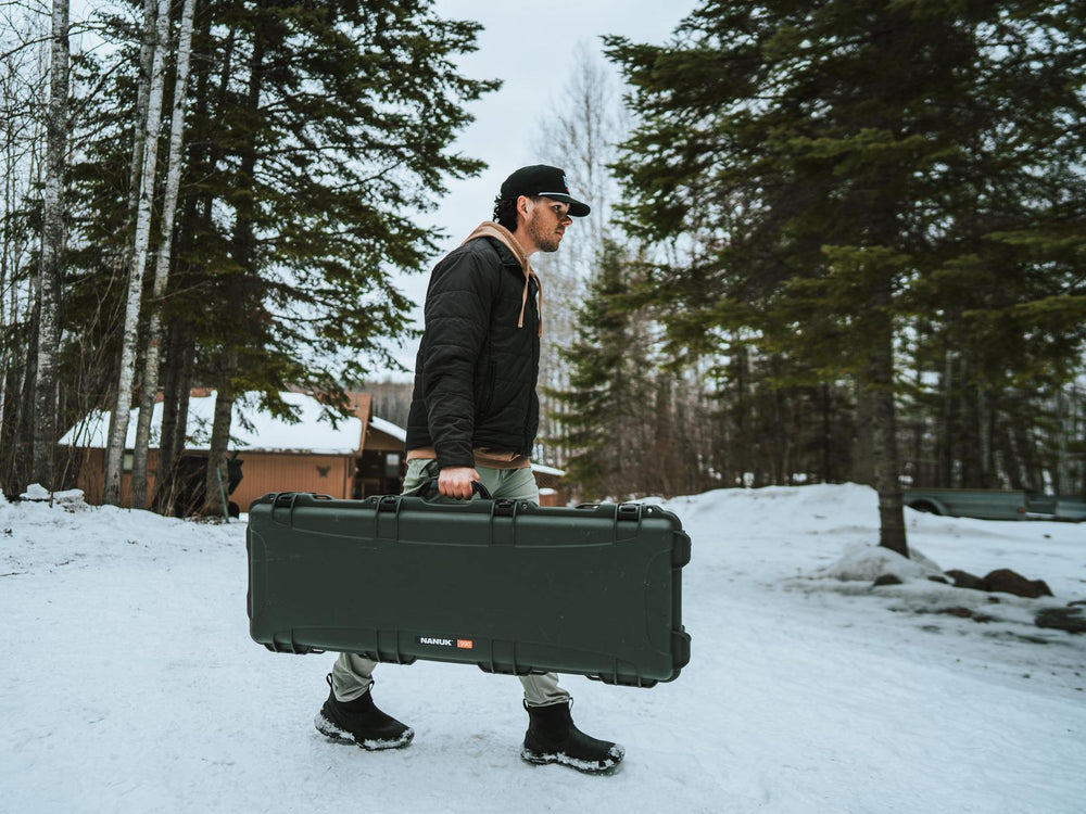 Person carrying a large black case in a snowy landscape with trees and a cabin in the background.