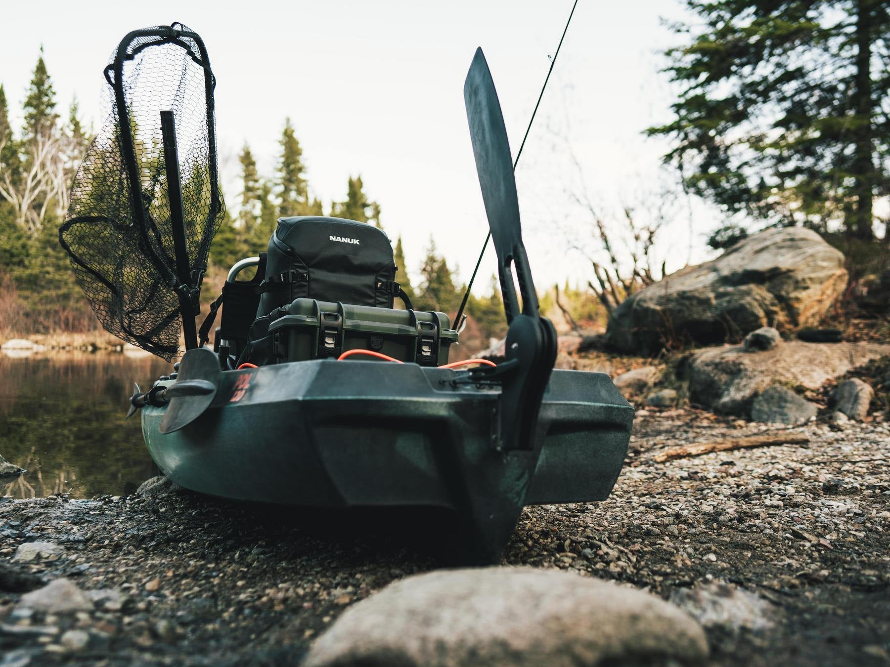 Fishing kayak with gear on a rocky shore near a body of water with trees in the background