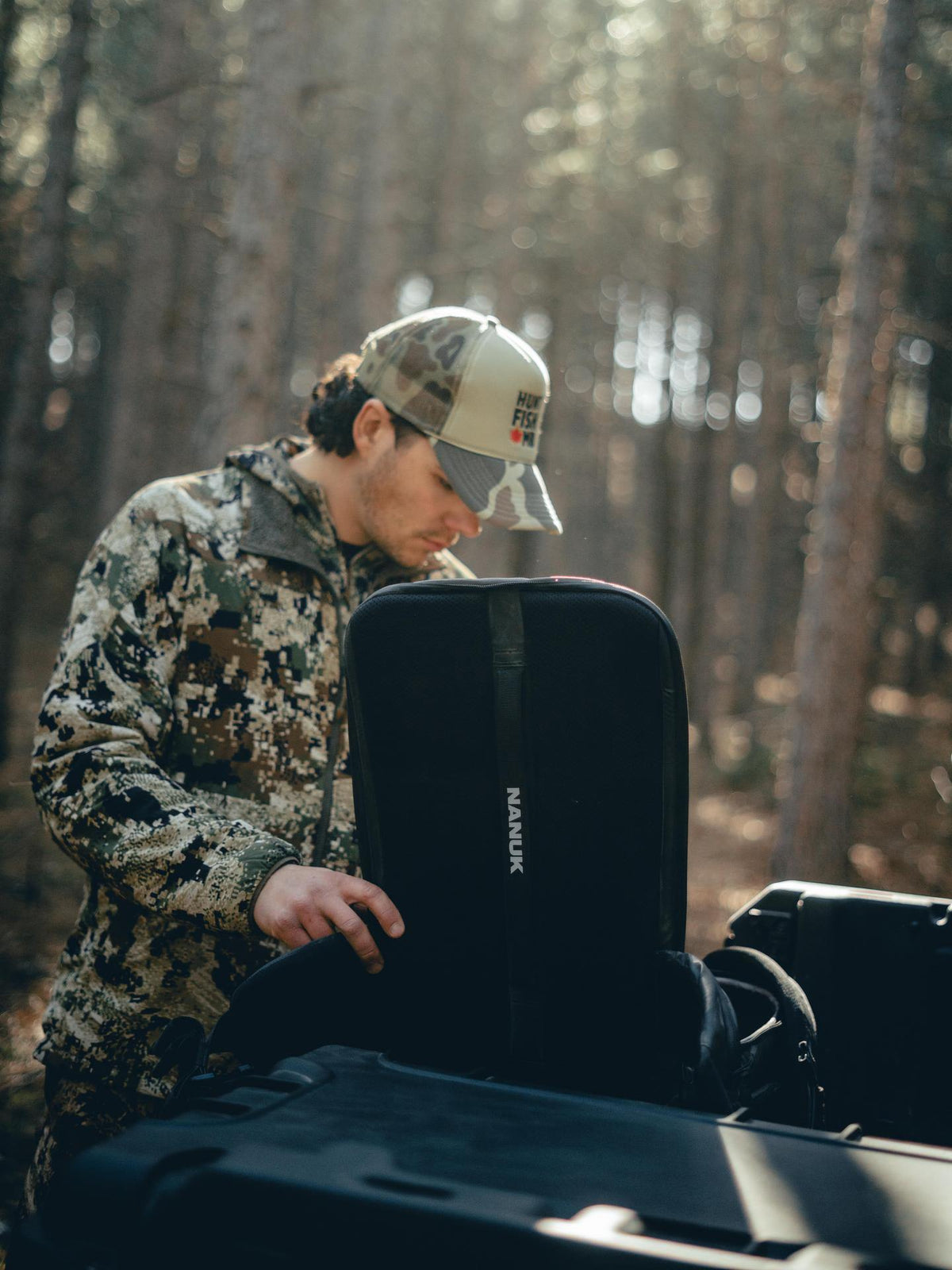 Man in camouflage outfit opening a black suitcase in a forest setting