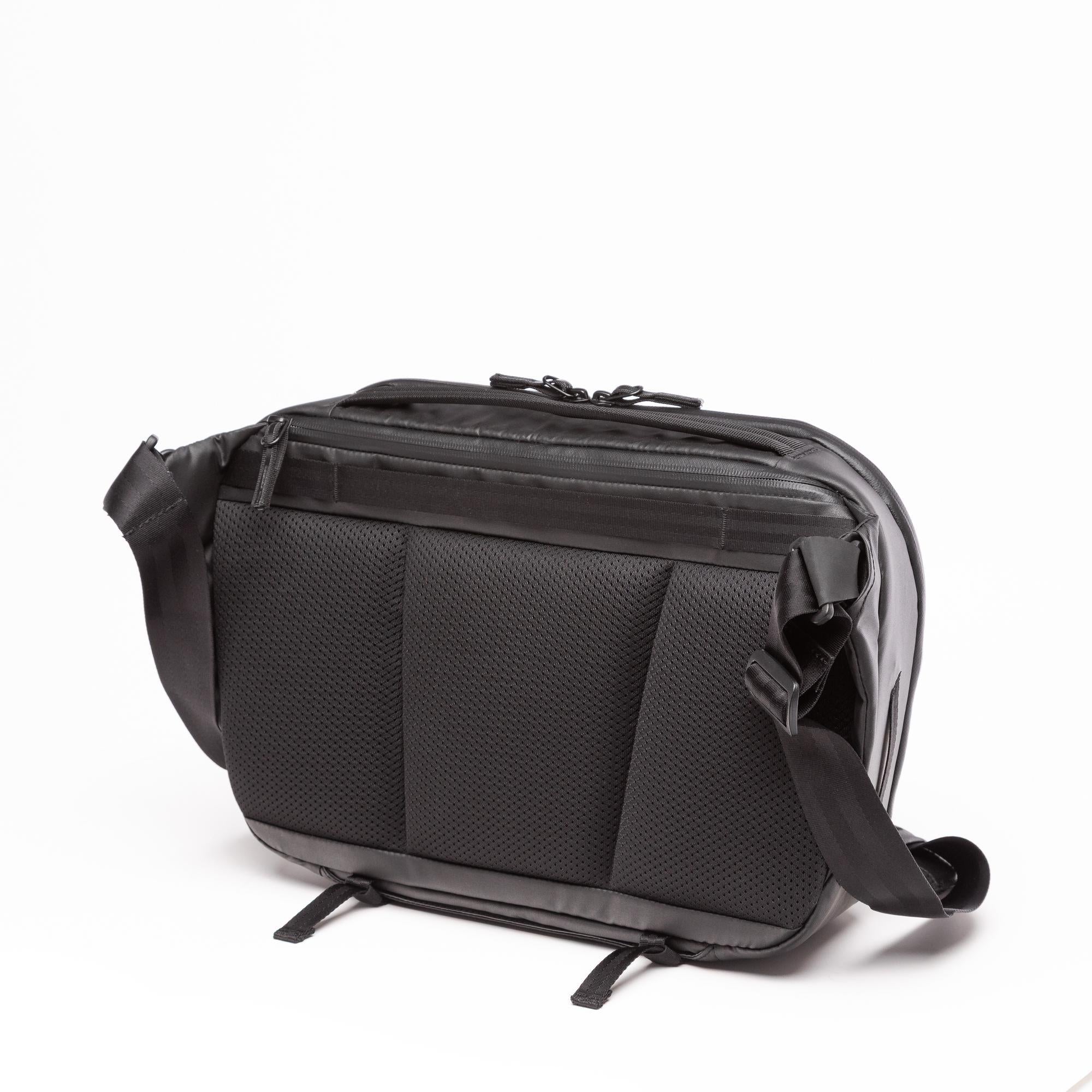 A black crossbody bag with a textured honeycomb pattern on the front pocket, a top zippered compartment, and an adjustable shoulder strap, shown on a white background.