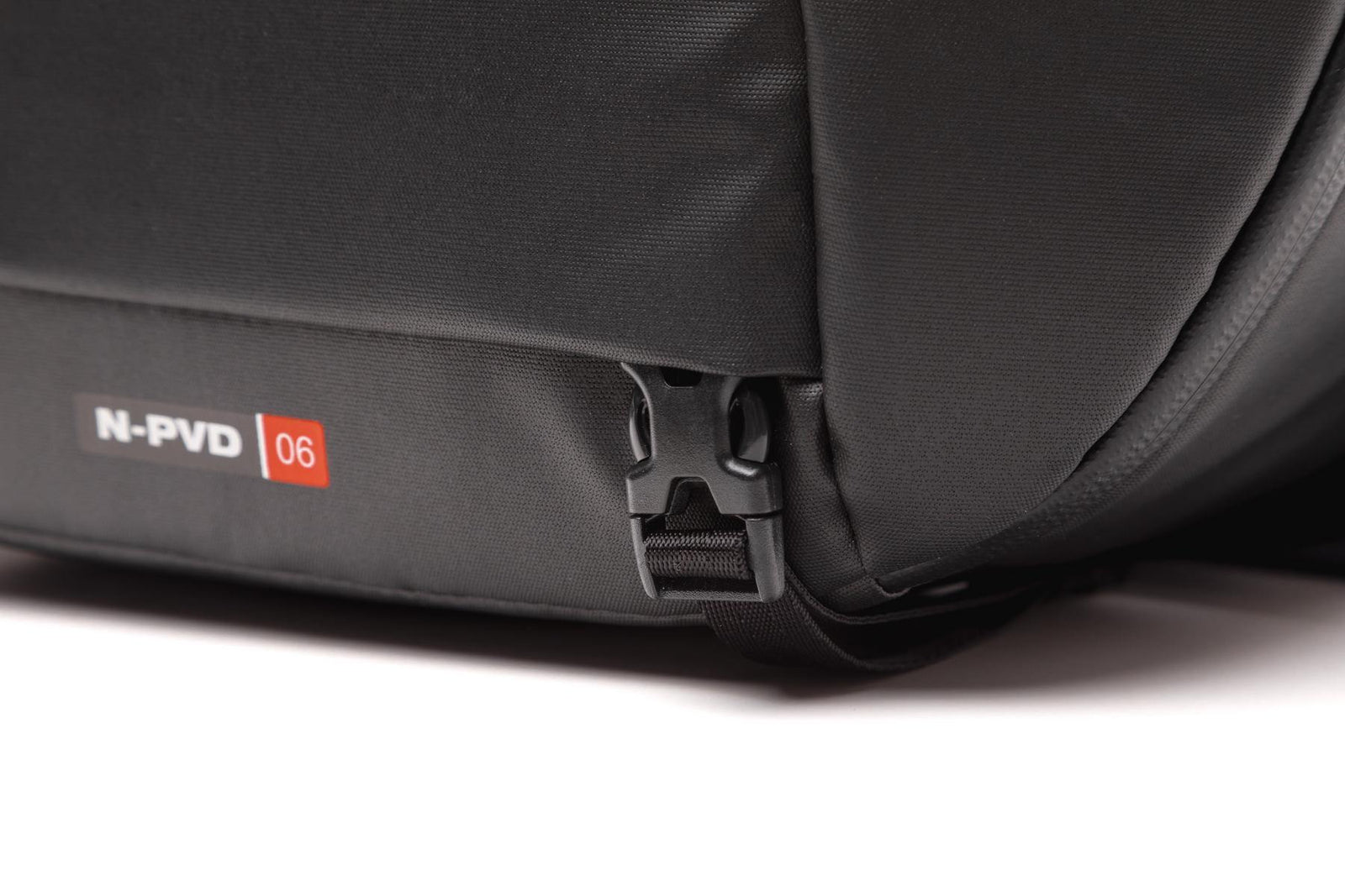 Close-up of a black nylon backpack showing the side seam, a black plastic zipper pull, and a small white and red 'N-PVD 06' label.
